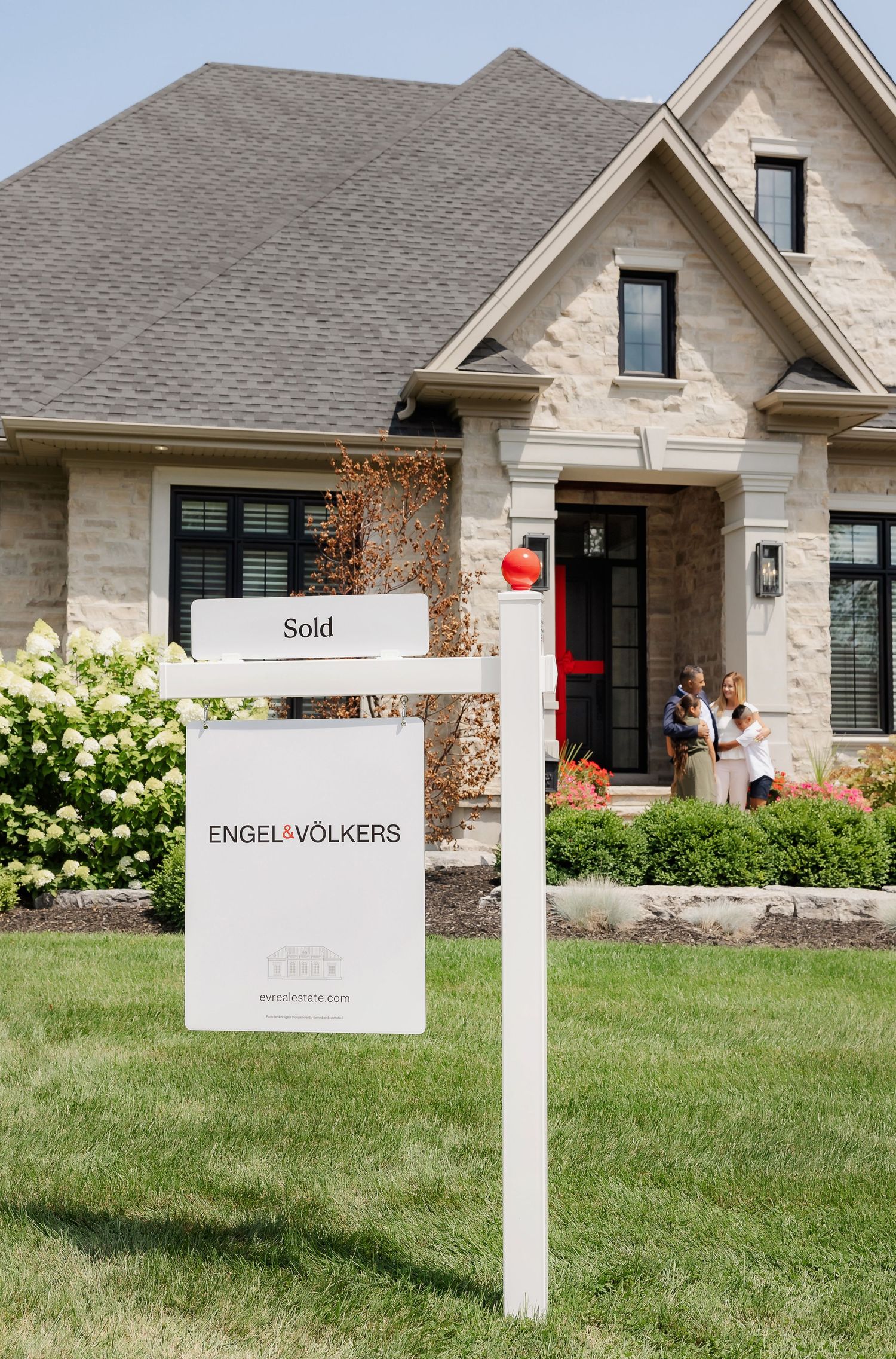 Real estate sold sign in front of modern stone house with manicured lawn and landscaping.
