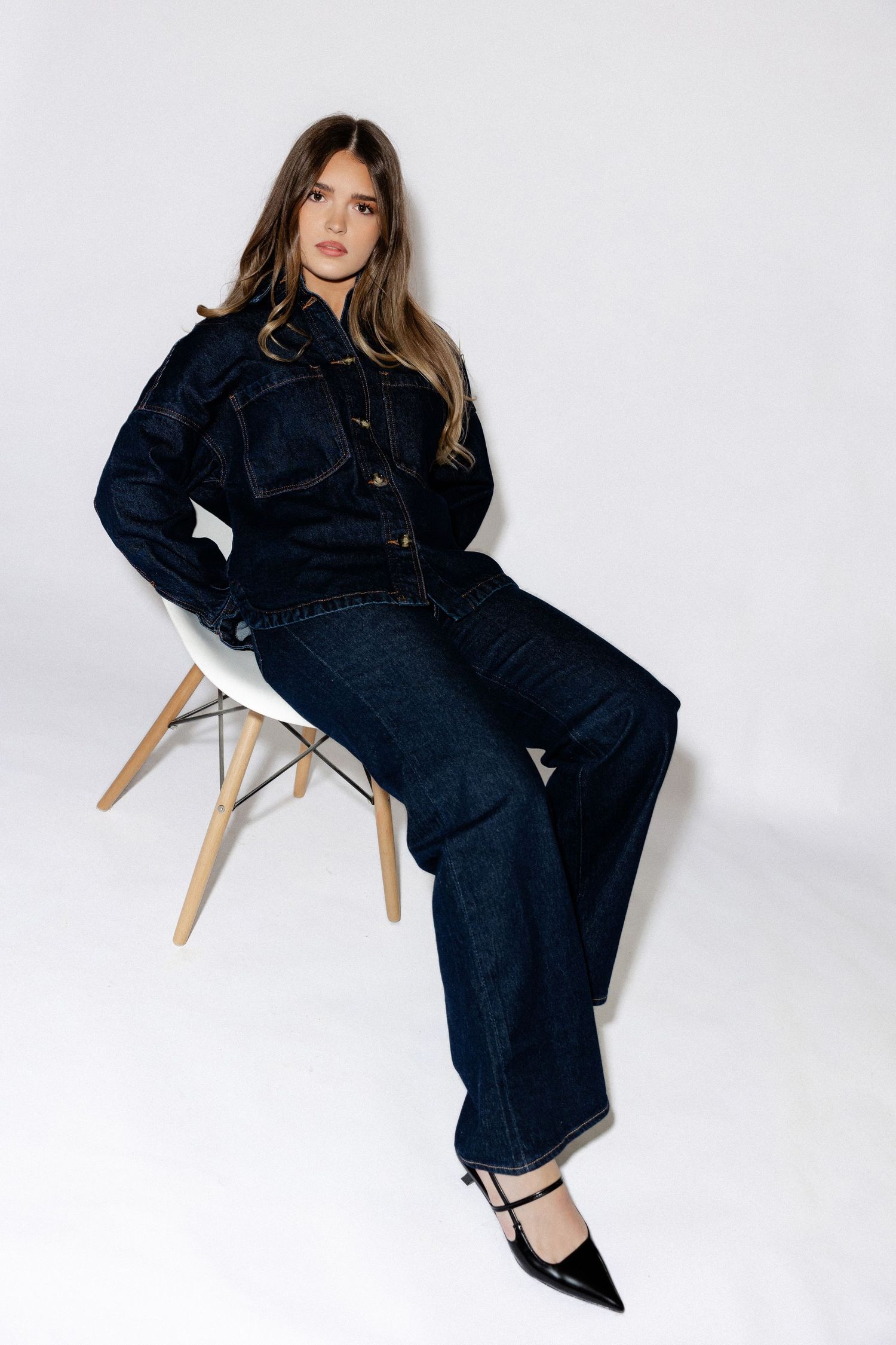 Modern fashion shoot featuring denim outfit and wooden chair against white backdrop.