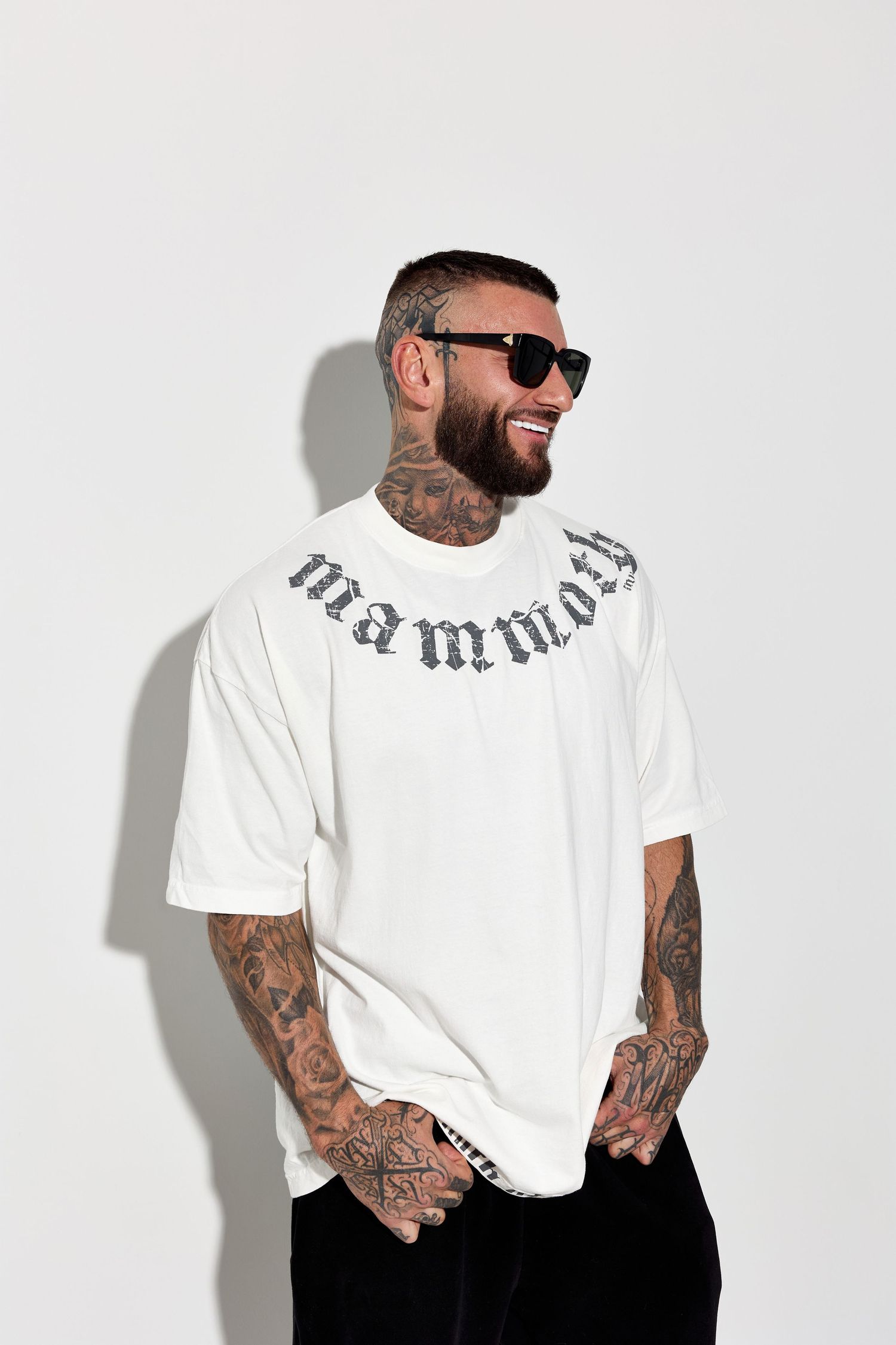Heavily tattooed model wearing white t-shirt and sunglasses against white background.