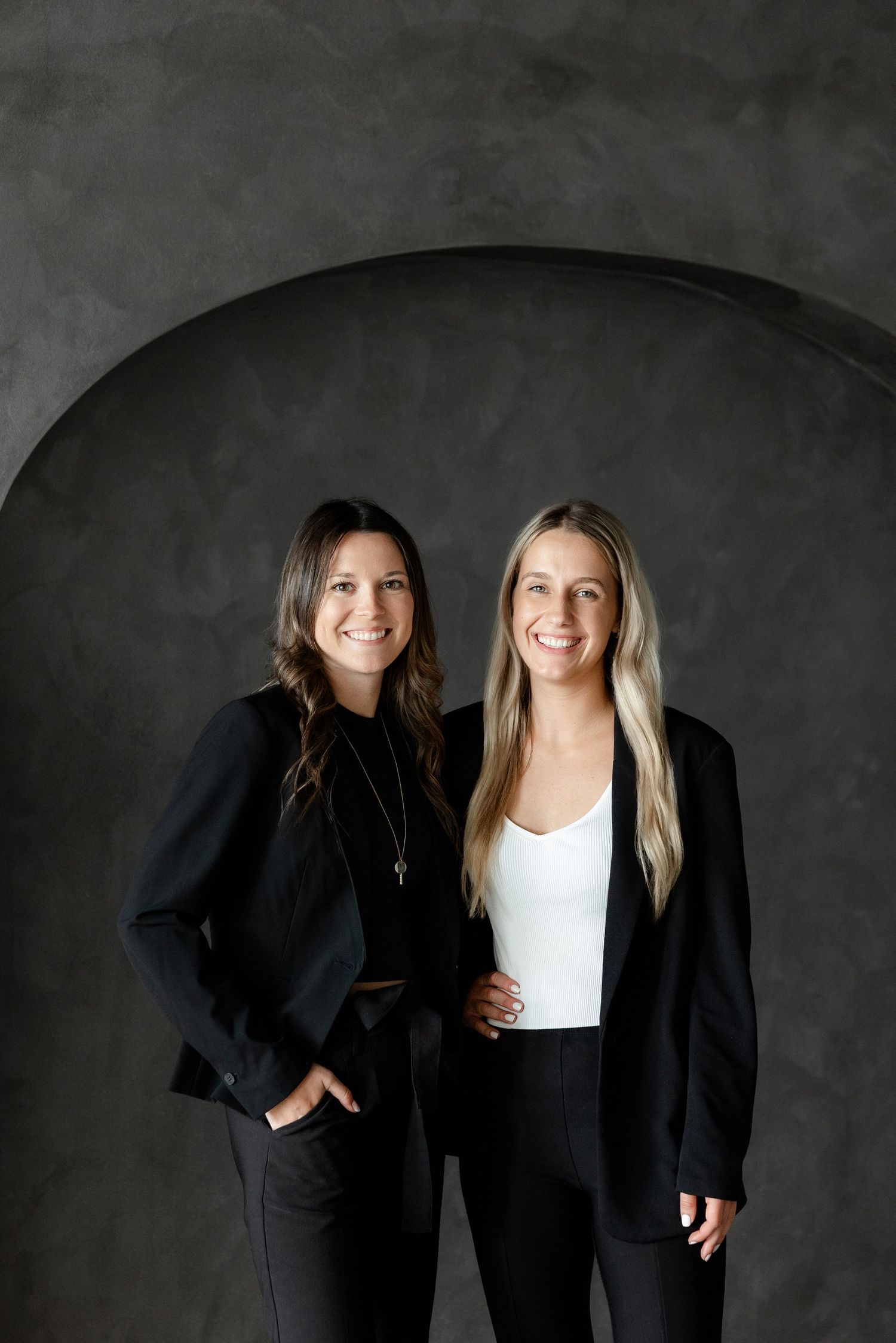 Two colleagues in business attire pose together against dark grey background.