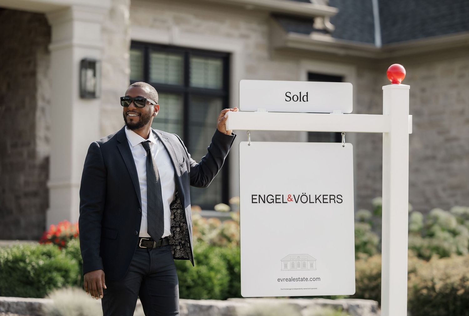Real estate agent in suit gestures towards Engel & Volkers sold sign in front of luxury home.