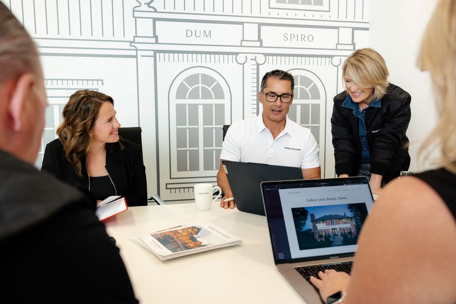 Business meeting with architectural drawings and digital presentations displayed.