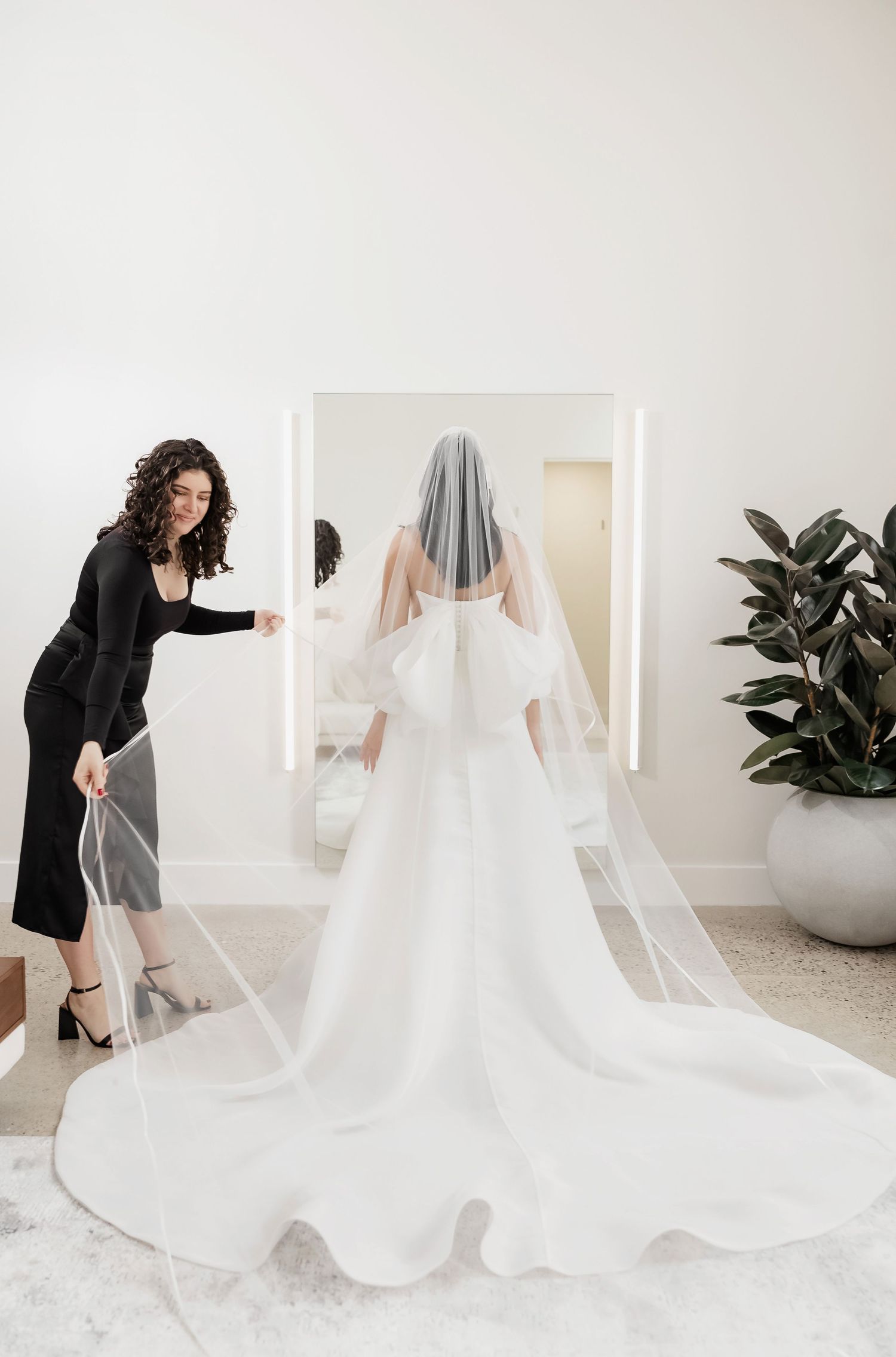 A wedding dress fitting showing the back view of a long train and veil being arranged by an assistant.
