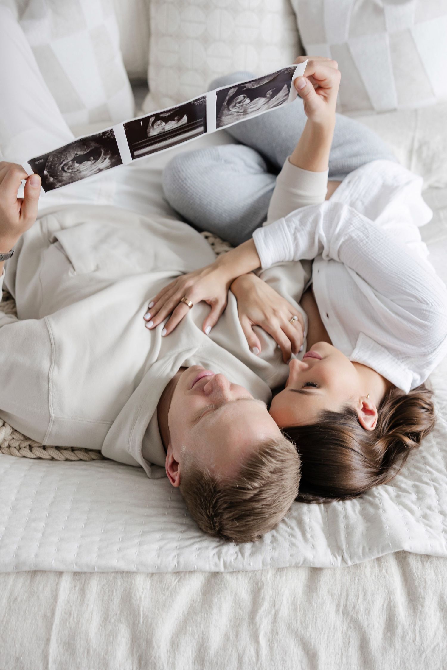 Couple lying on bed holding ultrasound photos in intimate pregnancy announcement scene.
