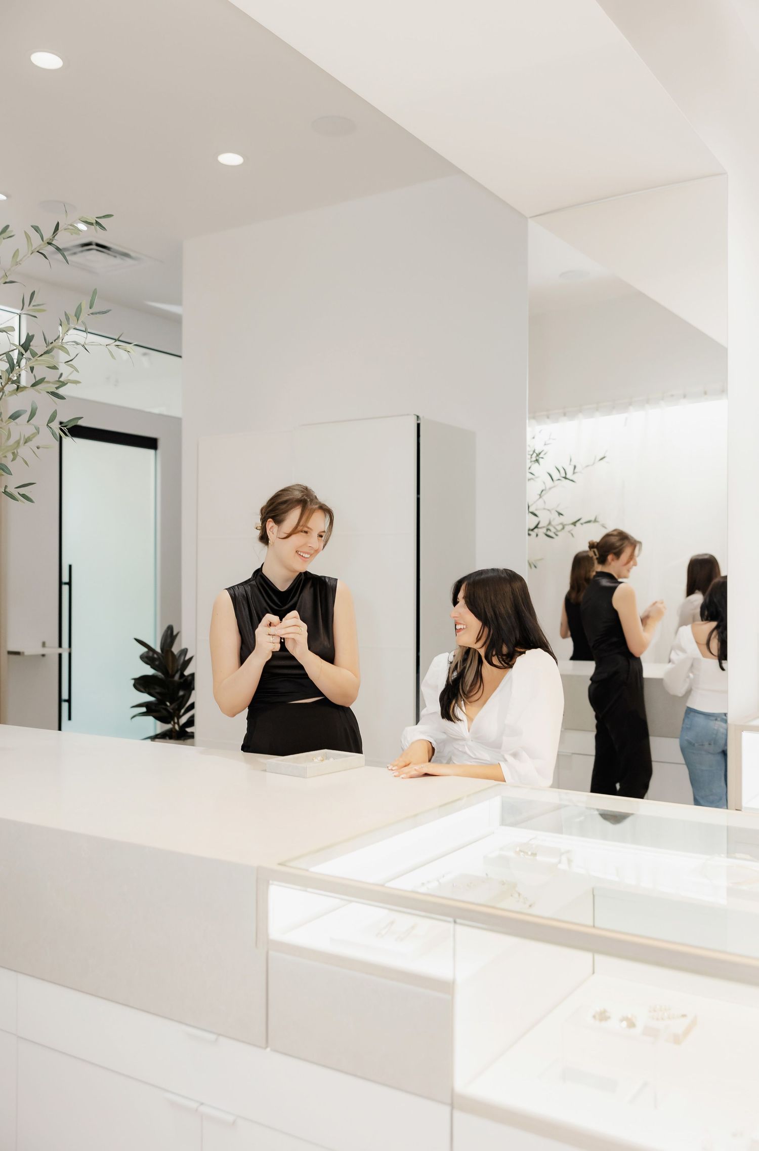 Customers interact at a bright white retail counter with mirrors and plant decor.
