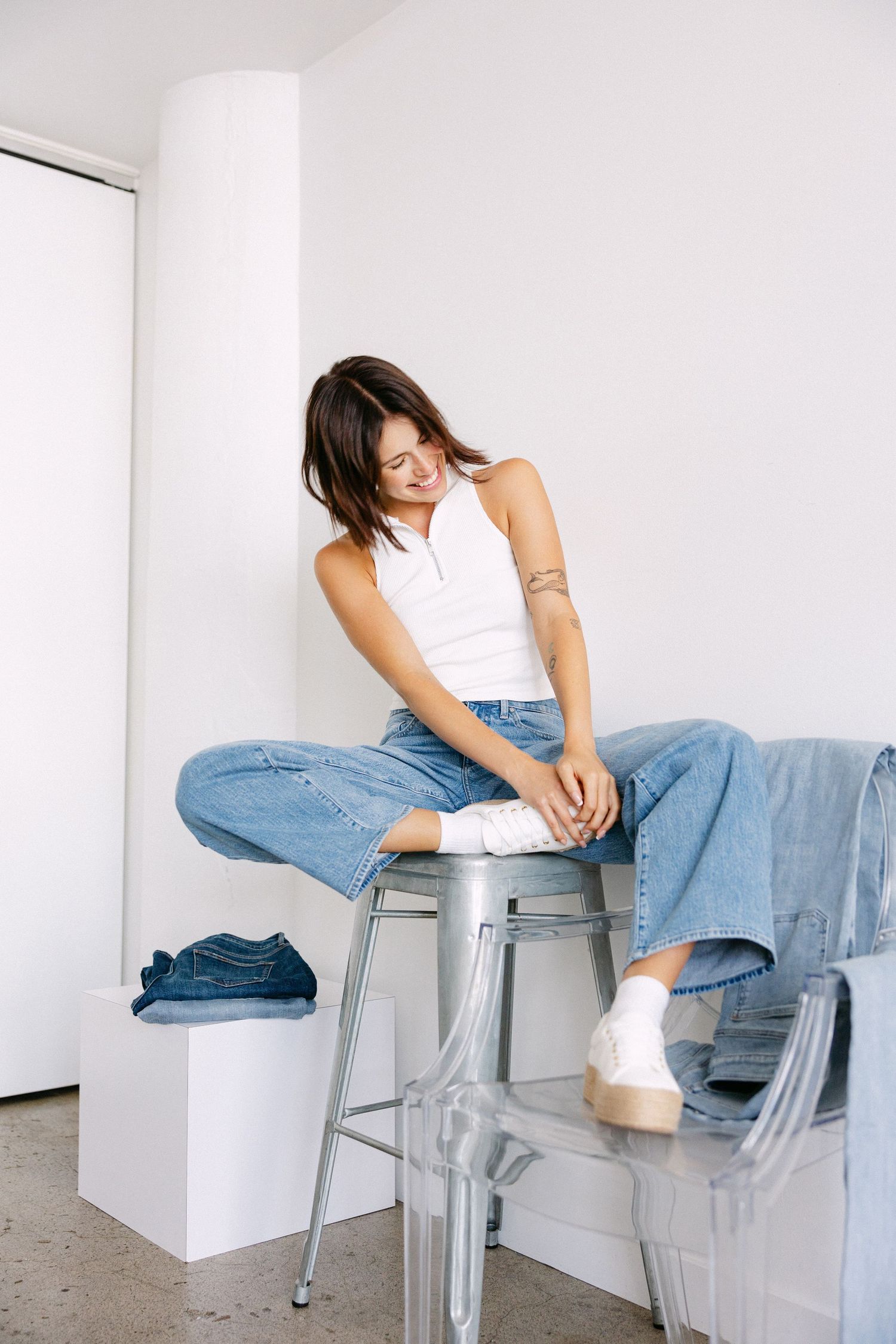 Editorial style photos showing relaxed poses on metal stool wearing casual denim and white top.