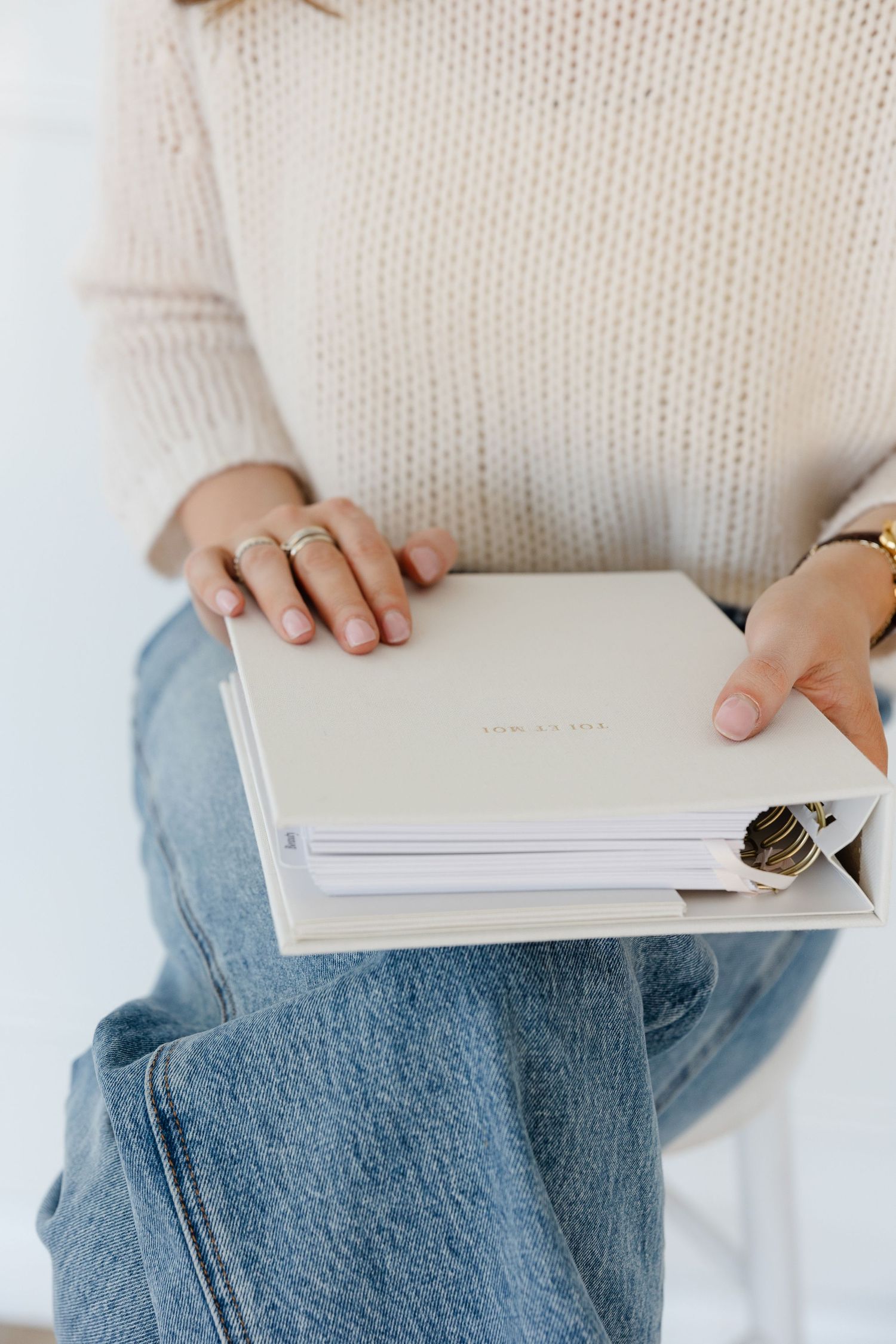Person in cream sweater holding white notebook while sitting in denim jeans.