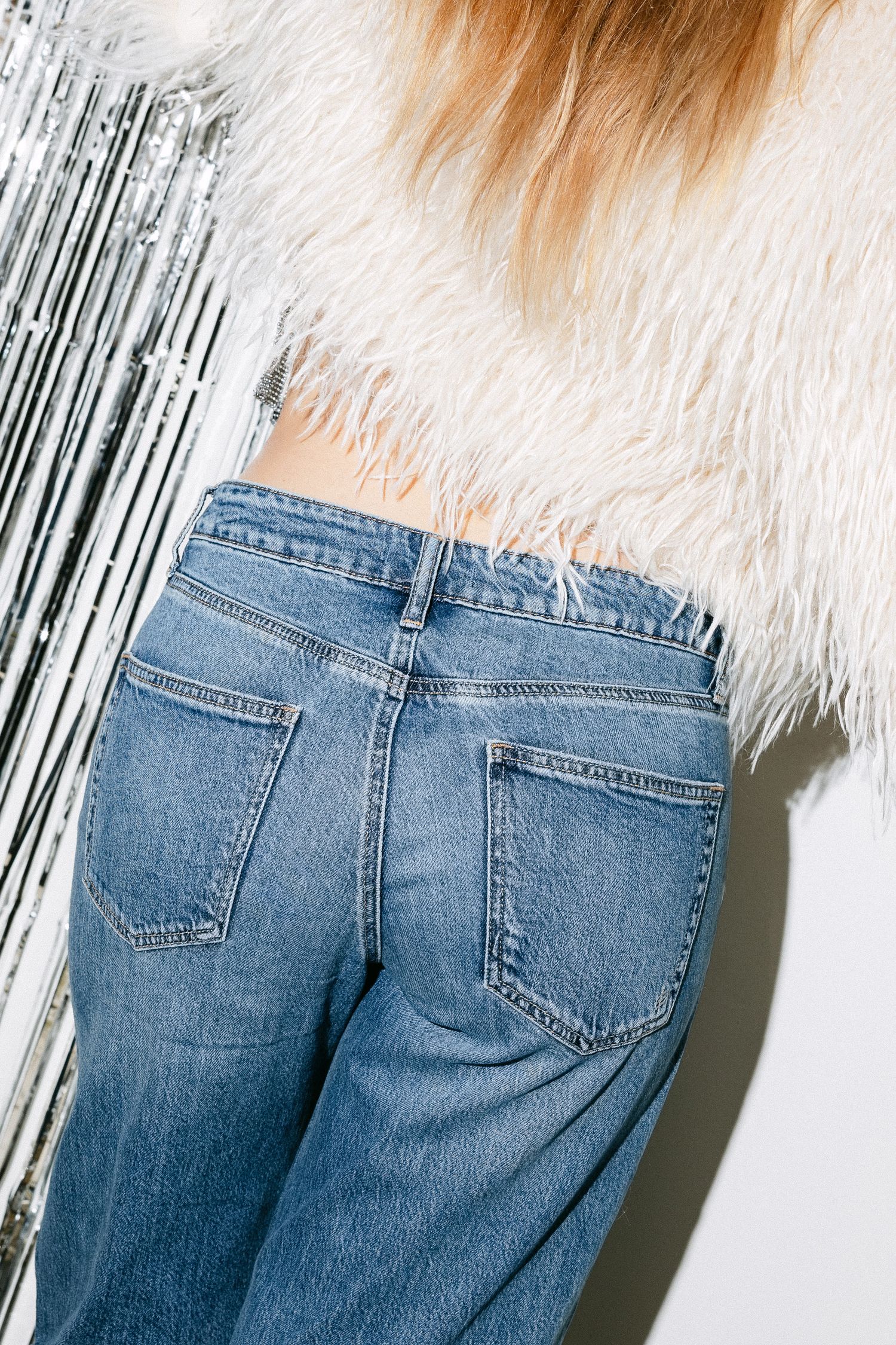 Close-up of denim jeans pocket detail with white furry jacket and metallic backdrop.