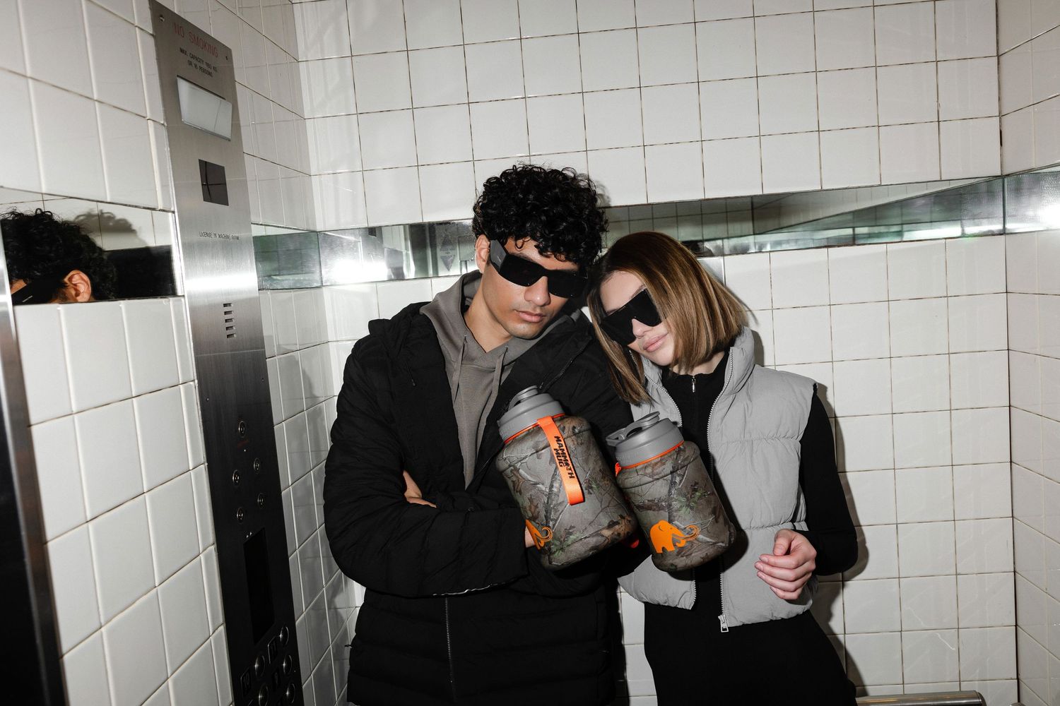 Two people in dark clothing lean against a white wall while holding beverages.