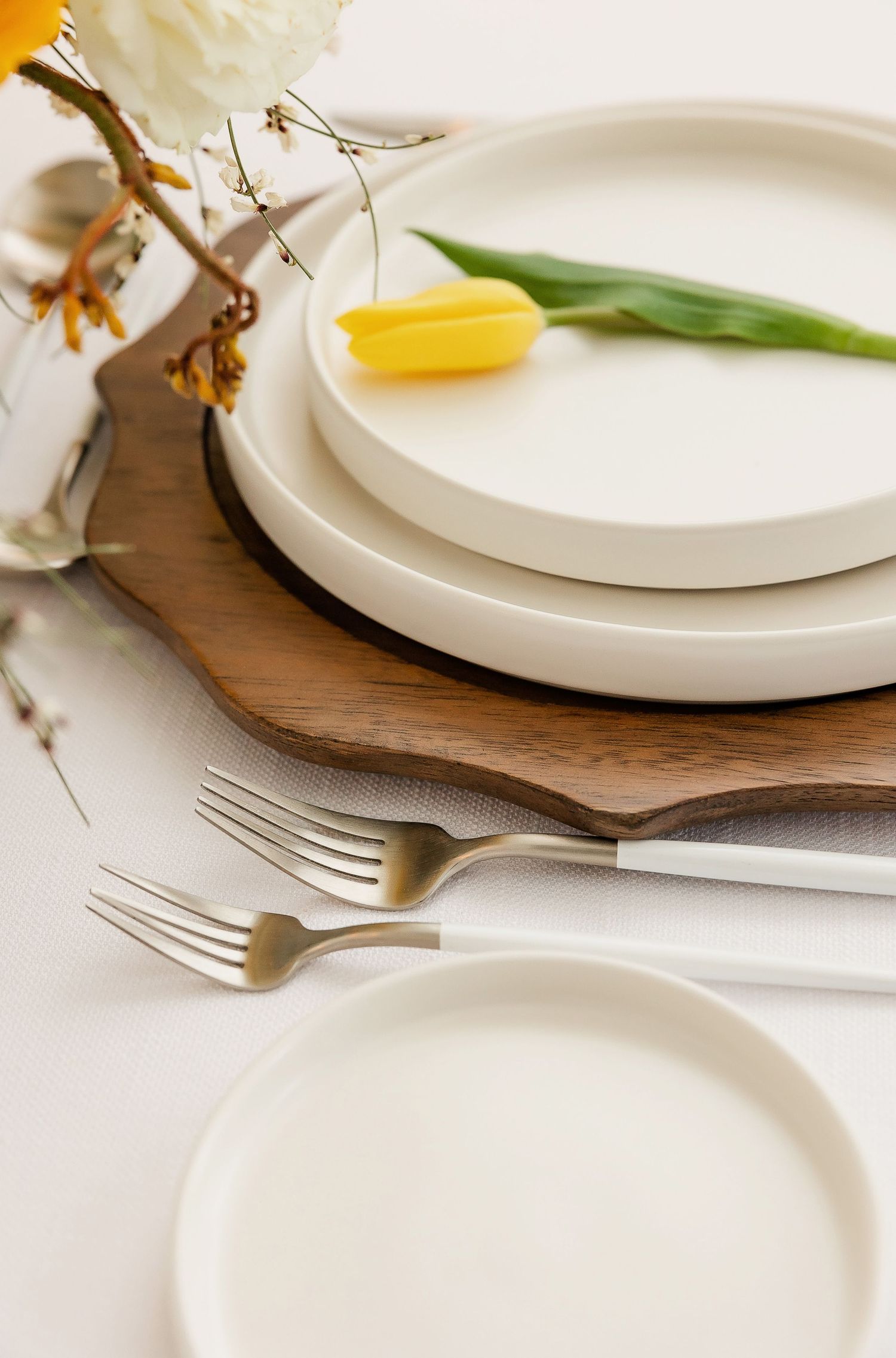 Minimalist white plates and gold utensils on wooden charger with floral accents.