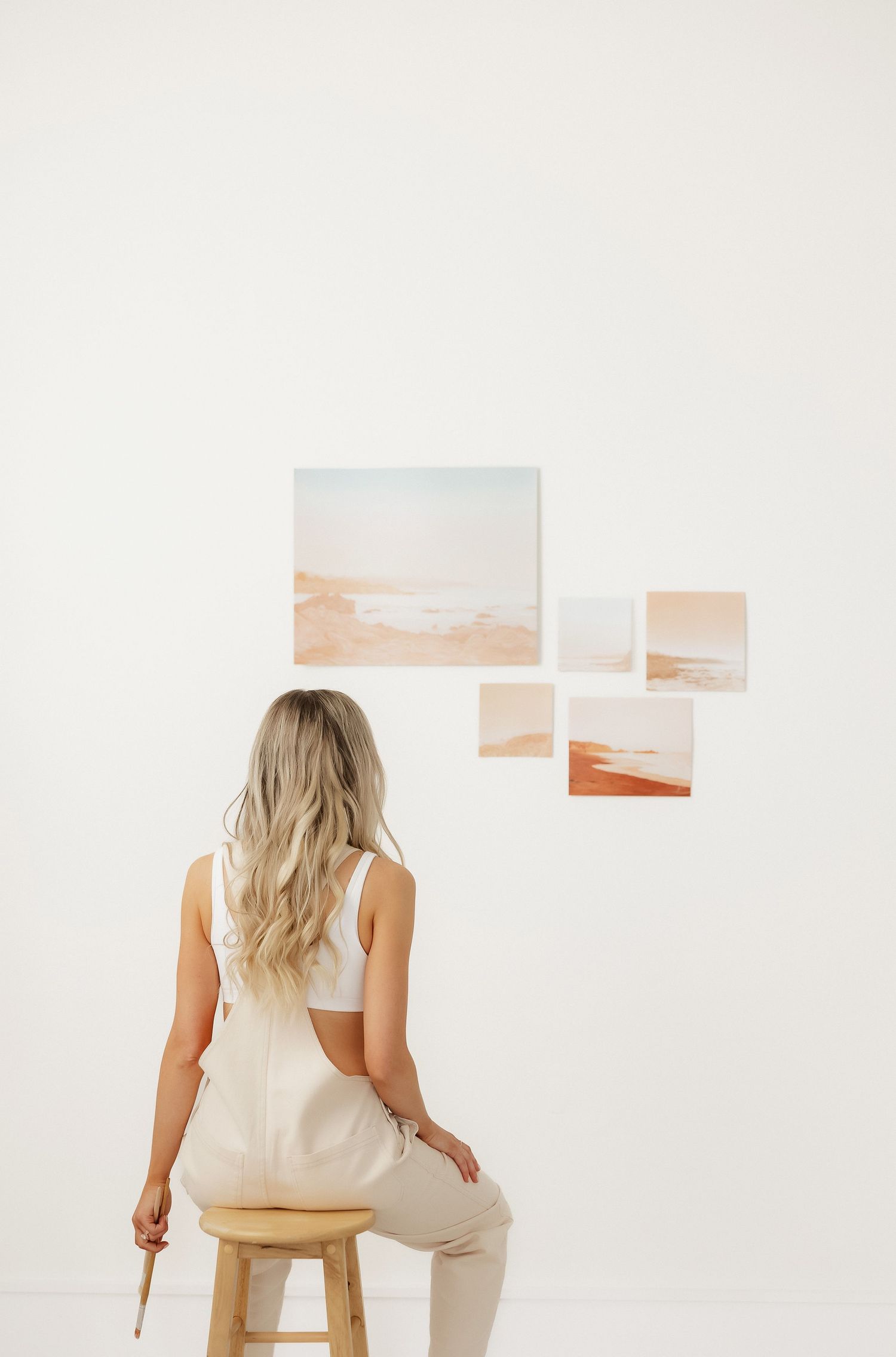 Artist in cream outfit sitting and viewing artwork displayed on white wall.