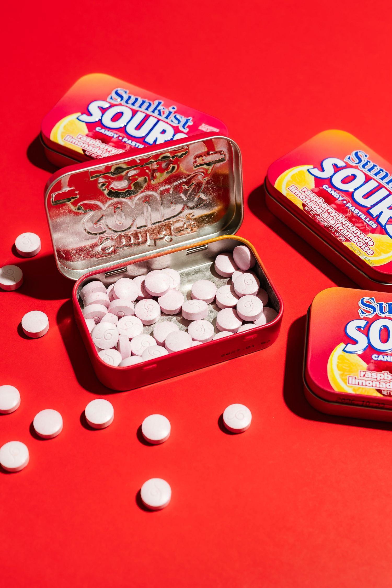 Sunkist Sours candy tins and white tablets arranged on bright red background.