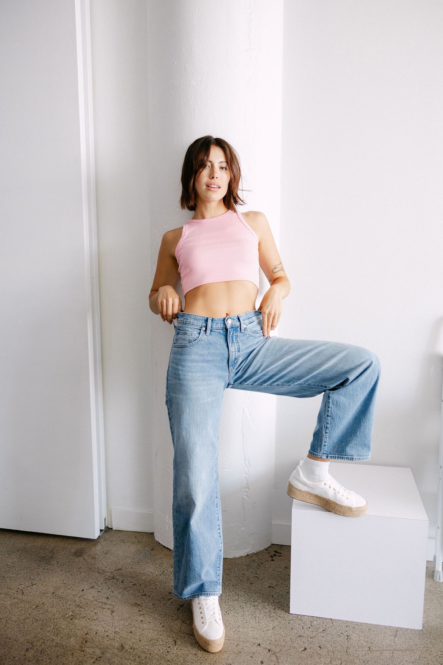 Model in pink crop top and baggy jeans poses casually against white backdrop with geometric white blocks.