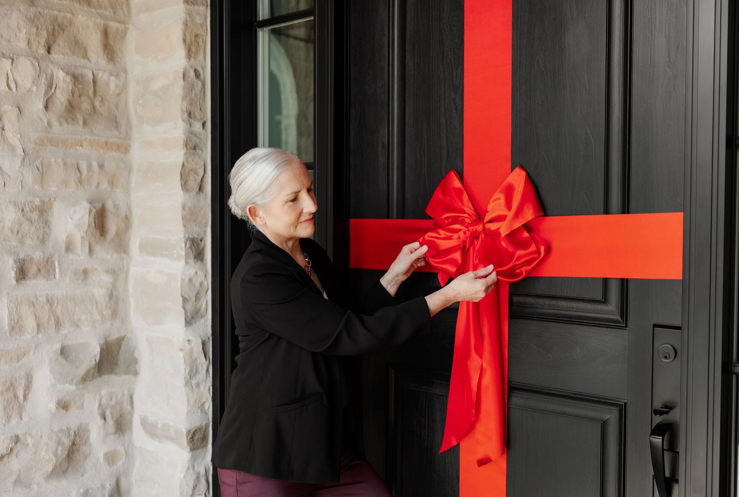 Person adjusts large red ribbon bow on black front door of stone house.