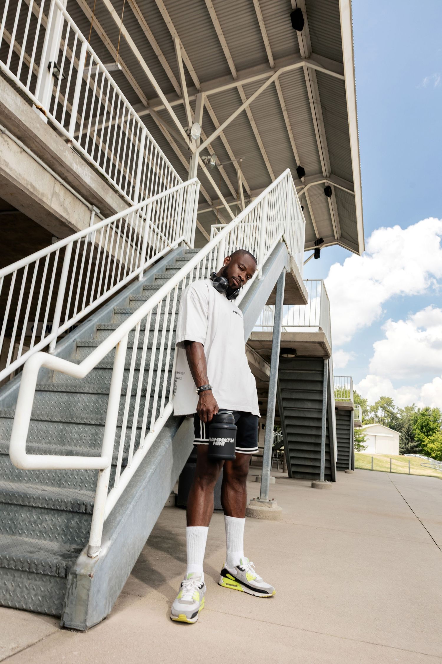 Athletic figure in white shirt and shorts posing near outdoor stairwell.