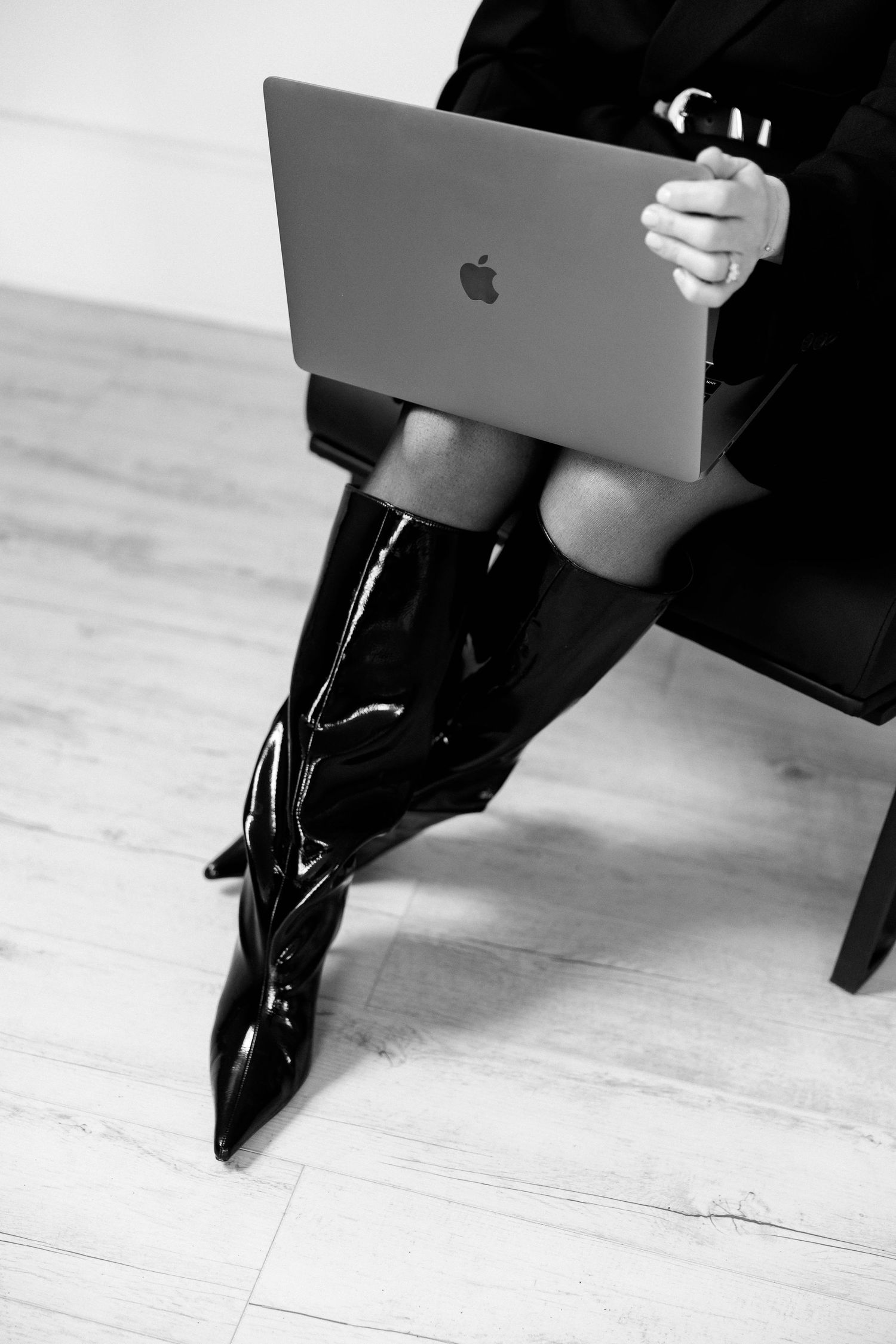 Black and white photo of MacBook and patent leather boots on wooden floor.