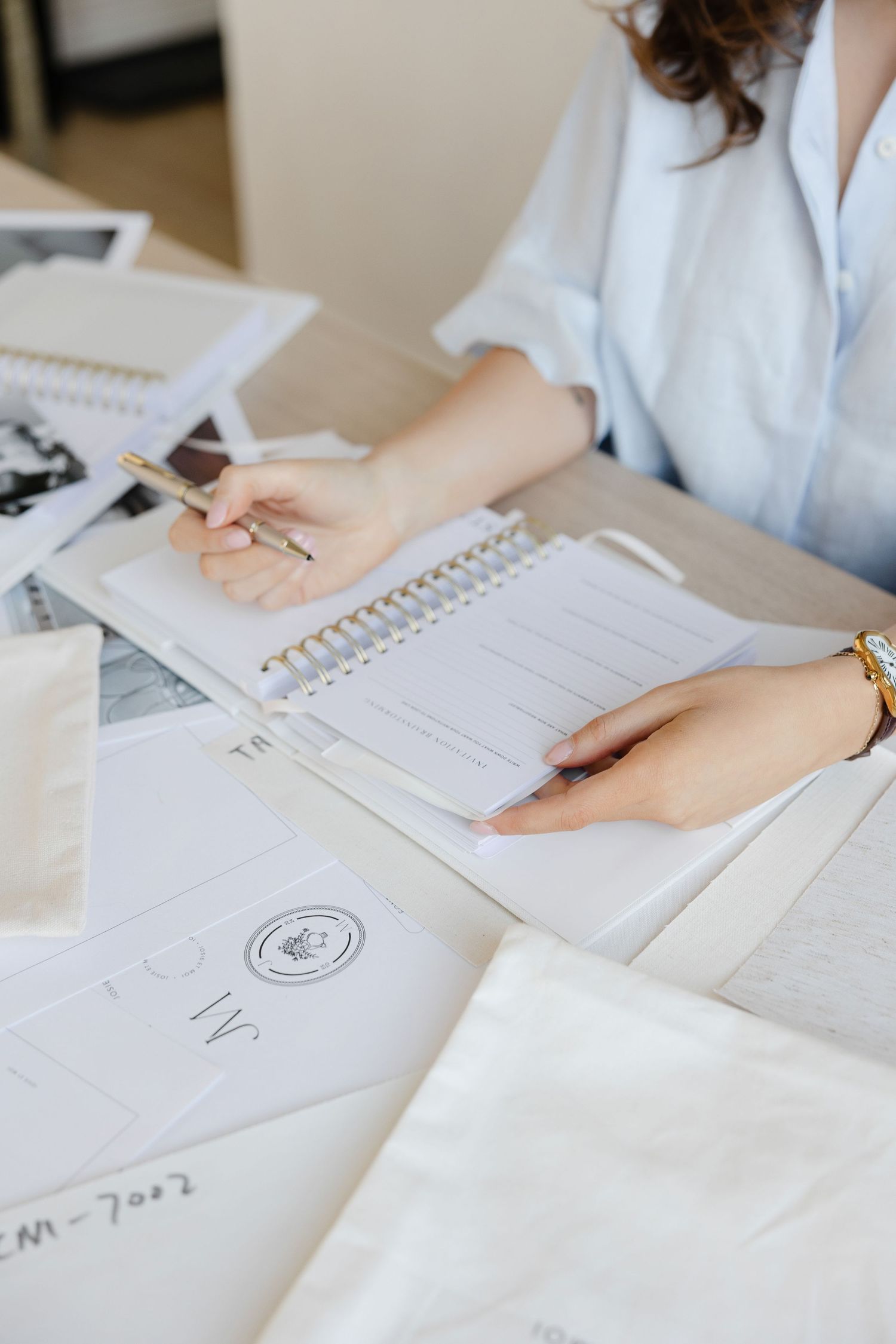 Hands writing in spiral notebook with branded materials and fabric pouches on desk.