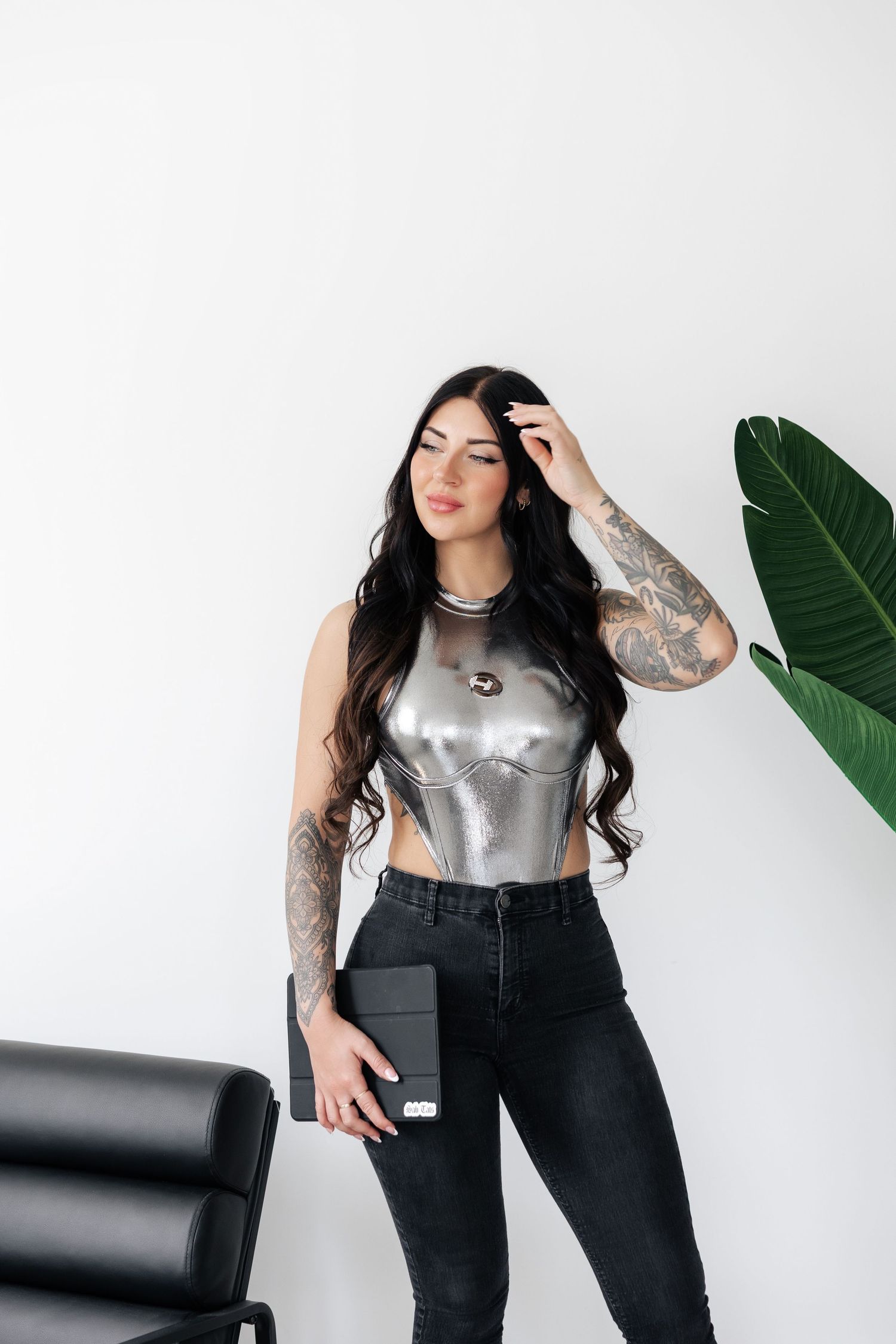 Model in silver metallic top and black jeans poses against white background with tropical plant.