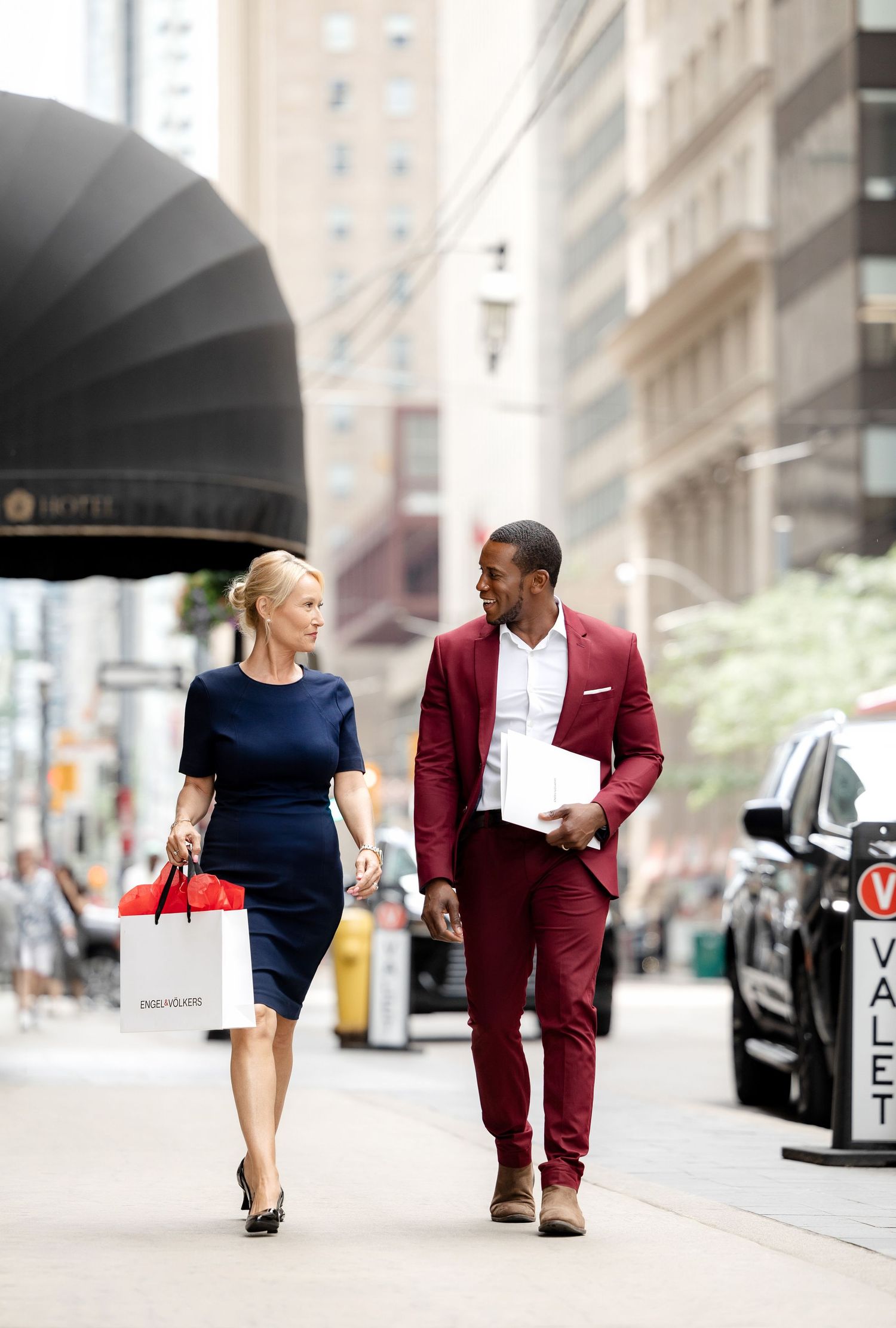 Two people walking down a city sidewalk past storefronts wearing business attire.