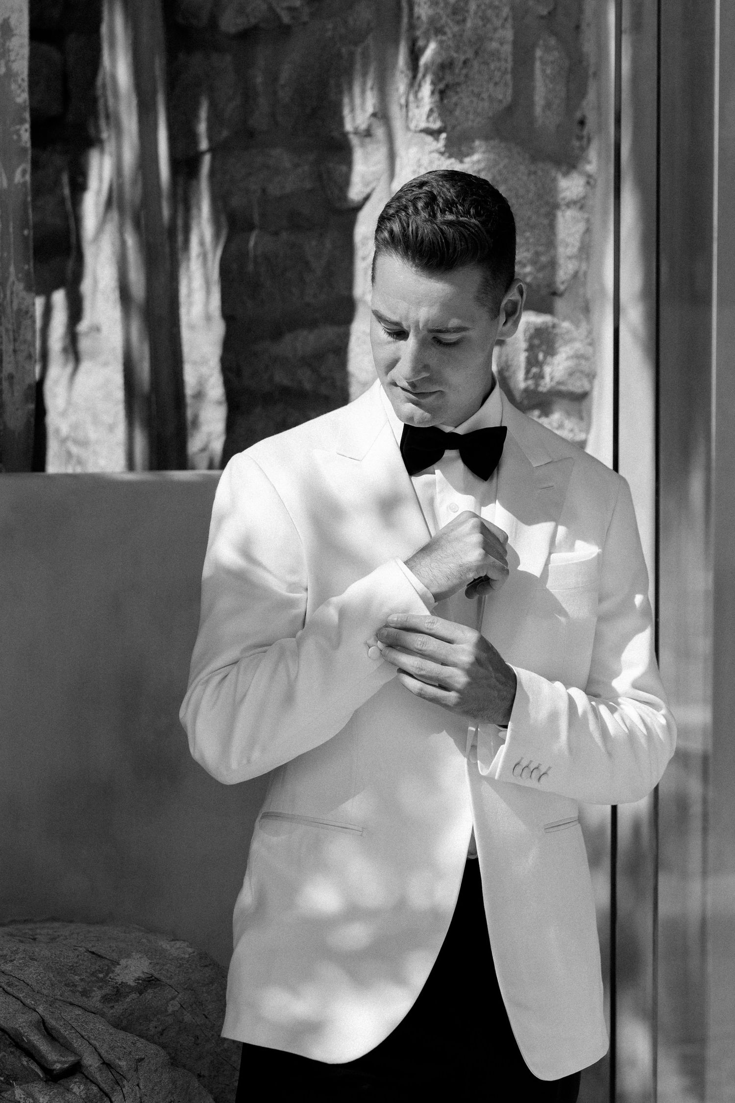 A groom in a white tuxedo jacket and black bow tie adjusts his cuffs in dramatic lighting.