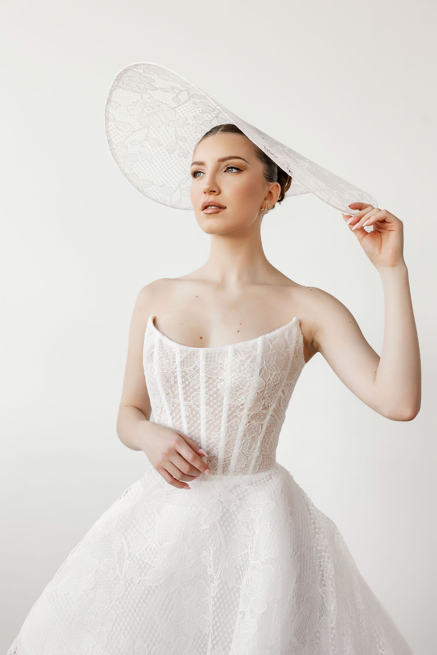 Bride wearing classic ball gown and wide-brimmed hat in minimalist studio setting.