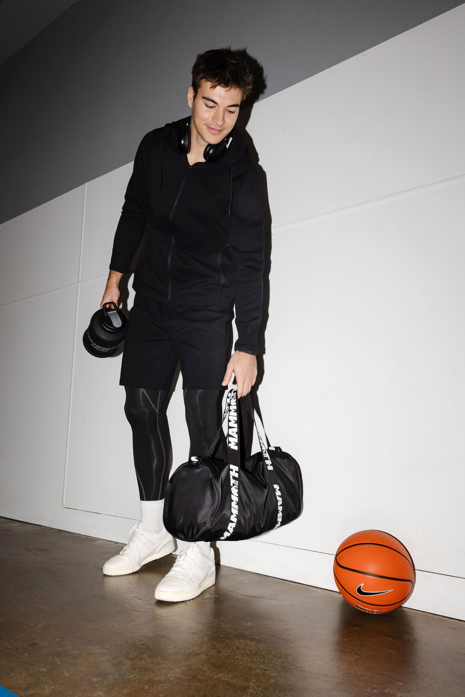 Person in black outfit sits with basketball and sports gear against white wall.
