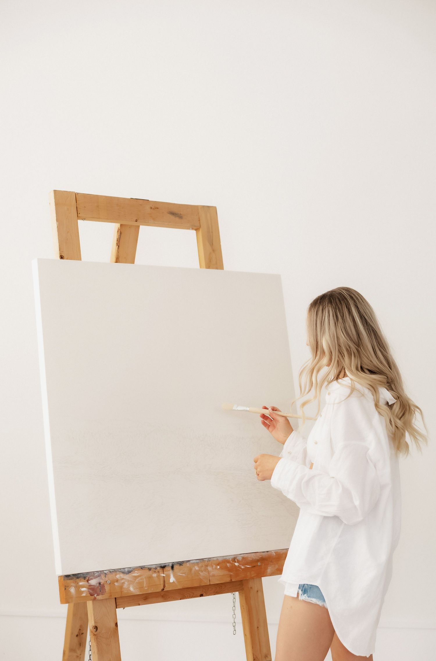 Artist in white shirt working at wooden easel with blank canvas against light background.