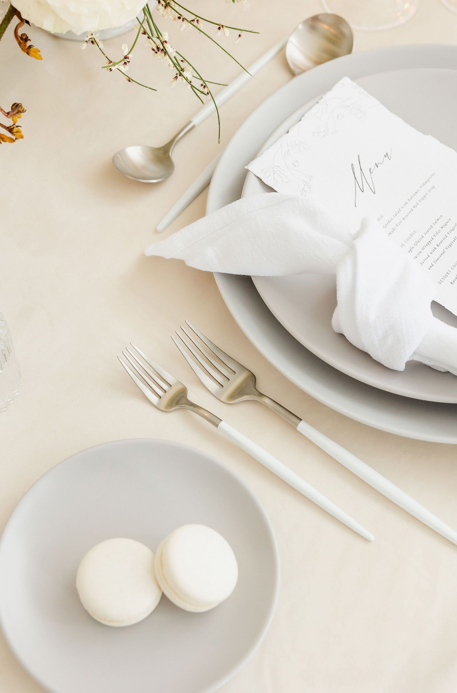White place setting with silver cutlery and macarons on a light beige tablecloth.