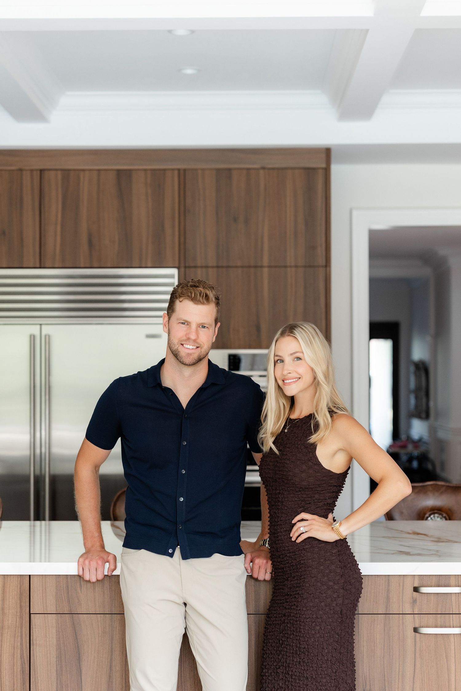A couple stands together in a modern kitchen with wood cabinets and stainless appliances.
