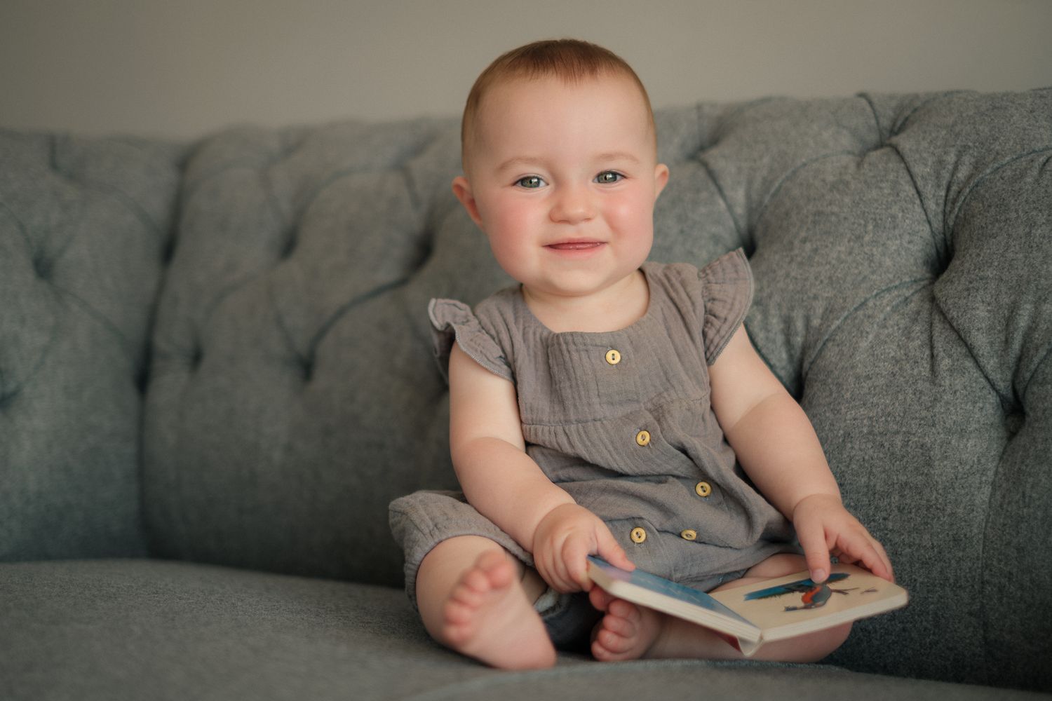 Baby smiling sat on a sofa holding book in natural light during an at-home photography session in Slaithwaite, Huddersfiled, West Yorkshire