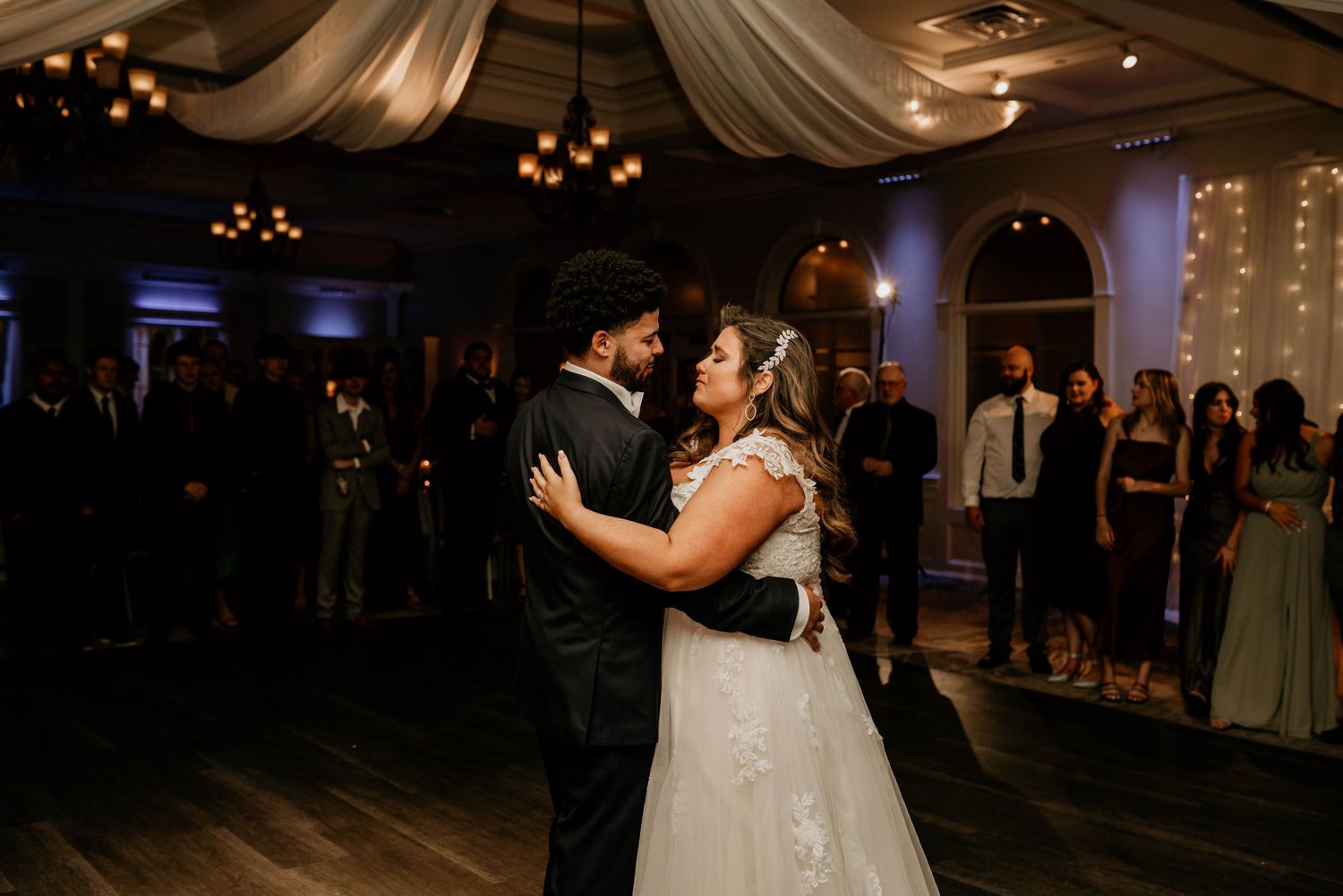 Bride and groom share their first dance at an elegant wedding reception with twinkle lights and draped fabric decor.