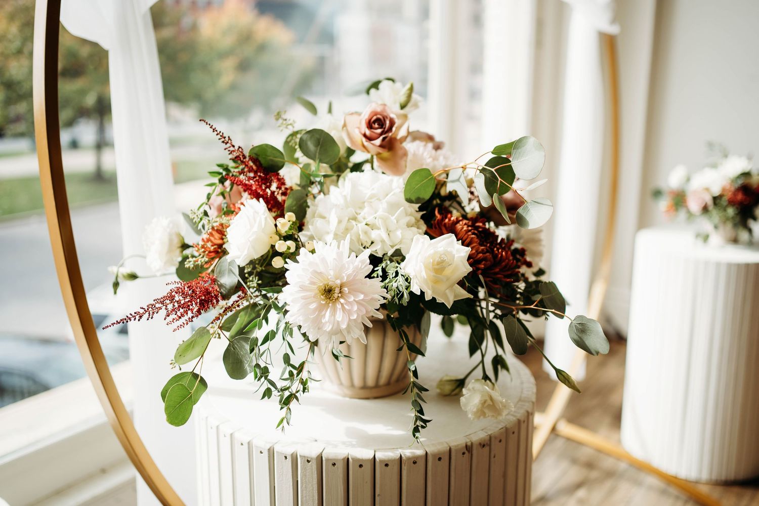 Wedding floral arrangement with white and burgundy blooms displayed near a window, photographed in soft natural light.
