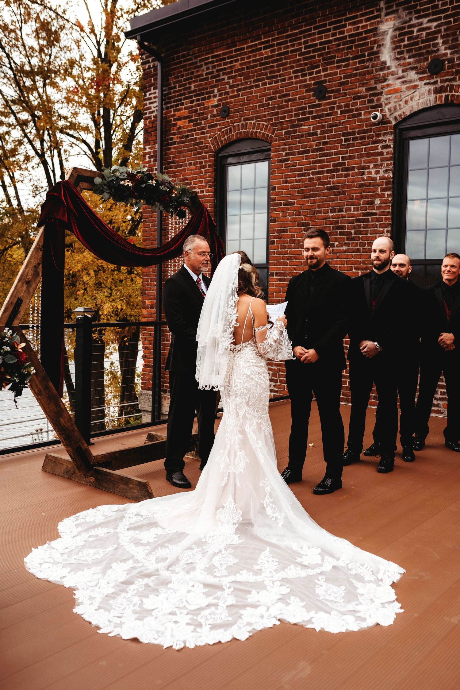 Wedding party in black suits hold up the bride's flowing lace train against brick building backdrop at sunset.