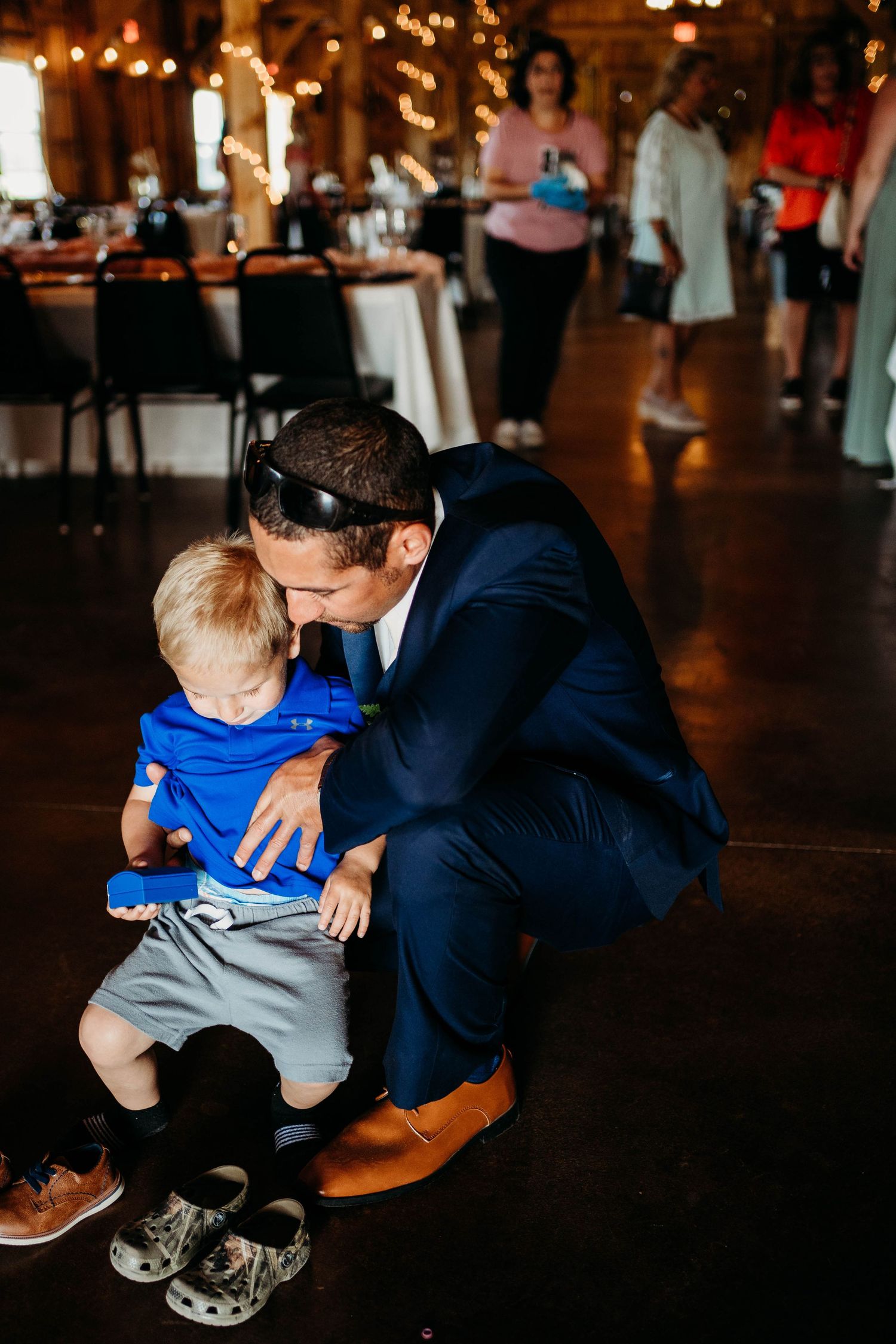 Groom in navy suit kneels beside toddler in blue shirt at evening reception venue with warm lighting.