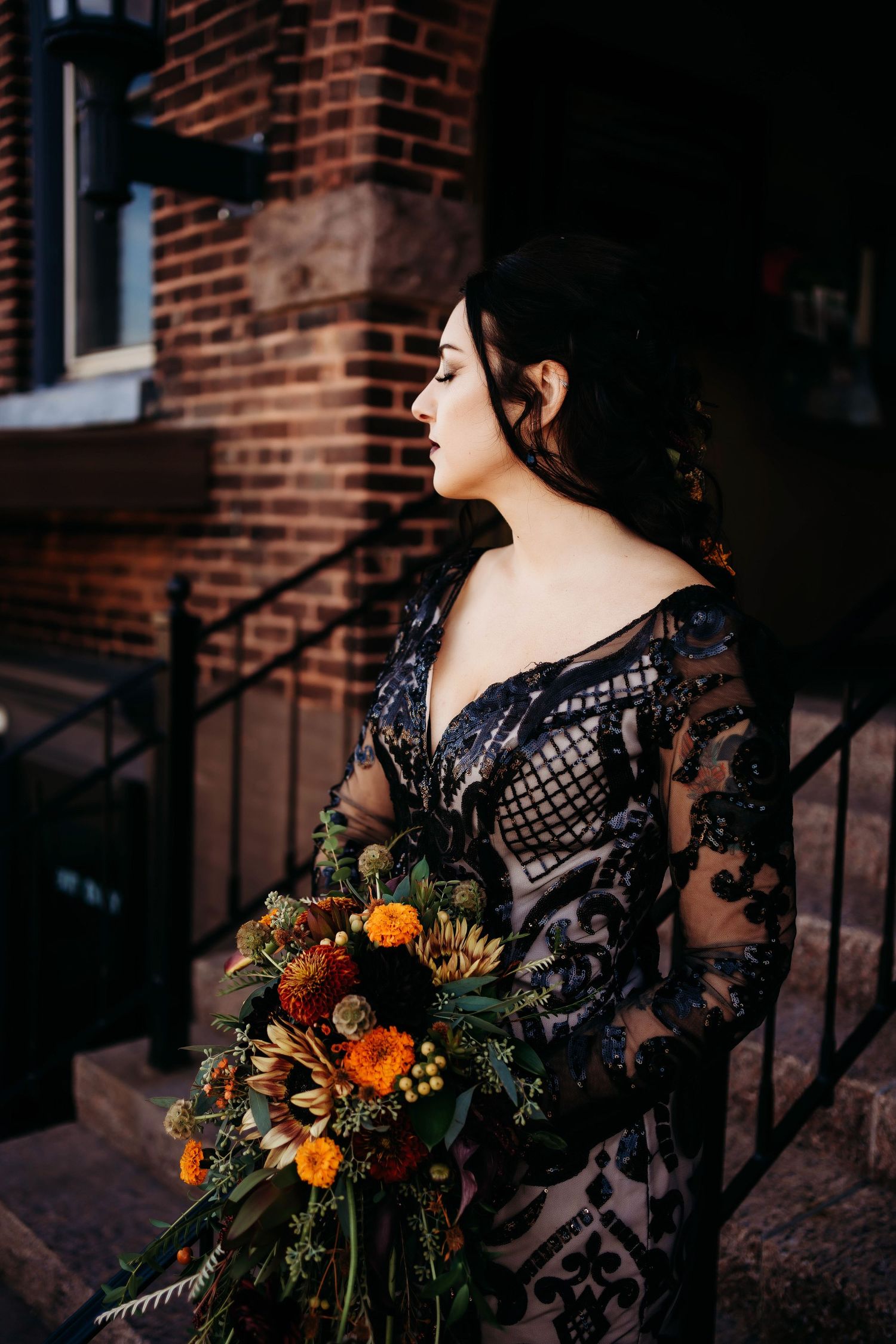 Editorial bridal portrait of a bride wearing a black lace gown and holding a bouquet against a brick wall.