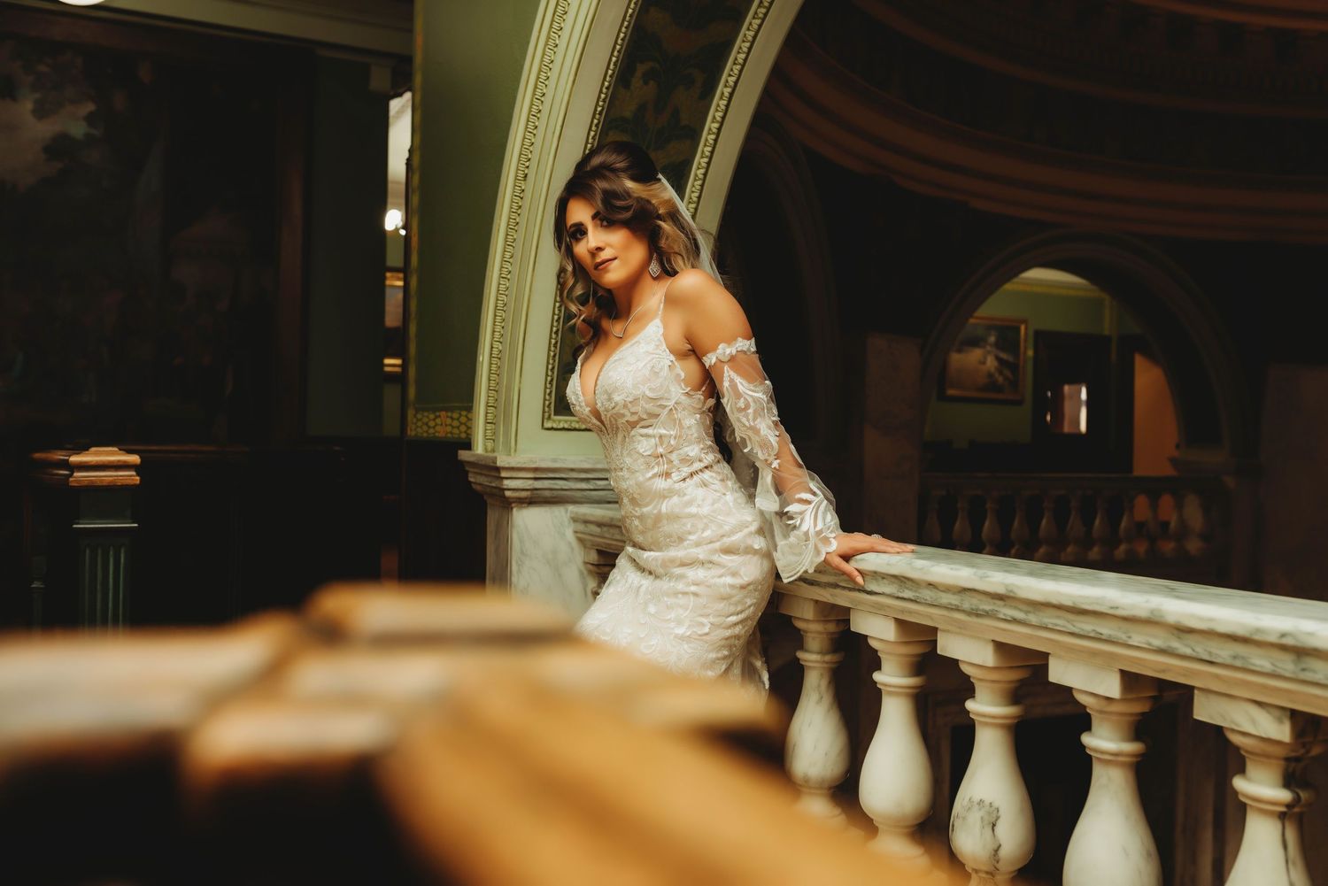 A bride in a white lace dress walks down a grand staircase in an elegant indoor venue.