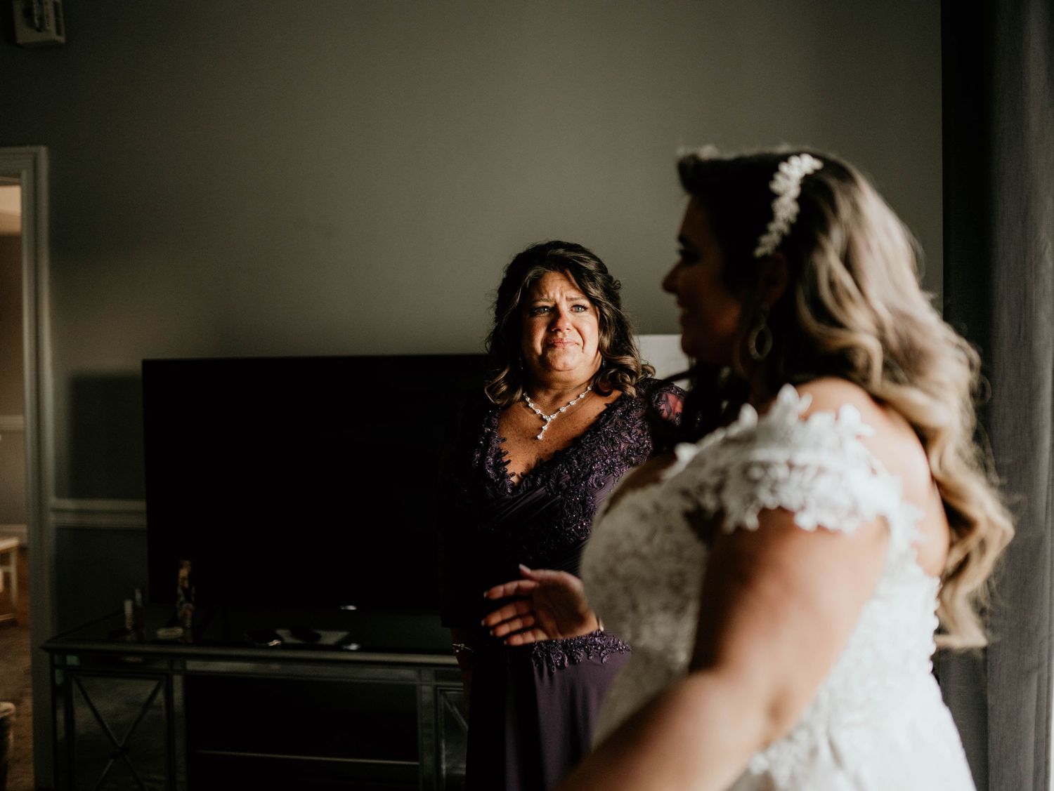 Bride and her mother share an intimate moment during wedding preparations in a dimly lit room.