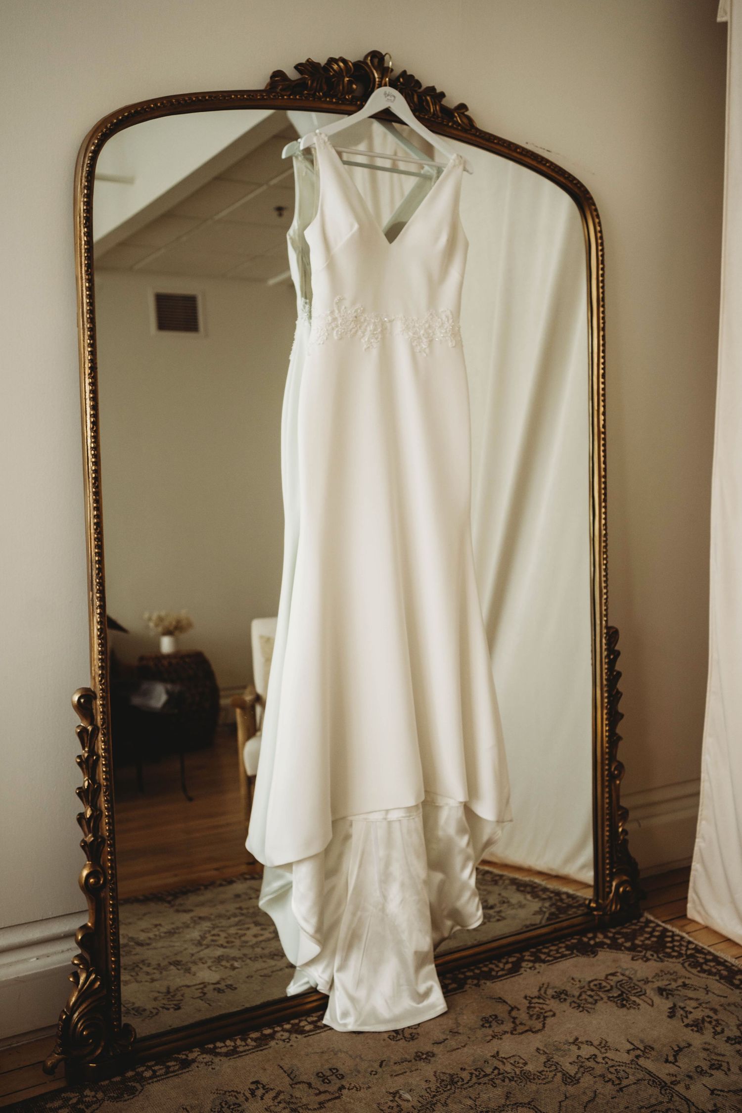 White wedding dress hanging in front of an ornate vintage gold mirror.