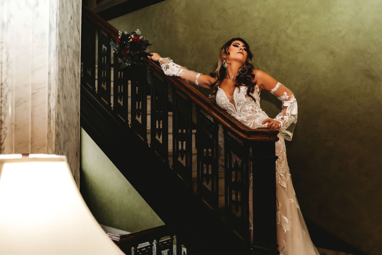 A bride in a flowing white wedding dress poses dramatically on a dark staircase with moody lighting.