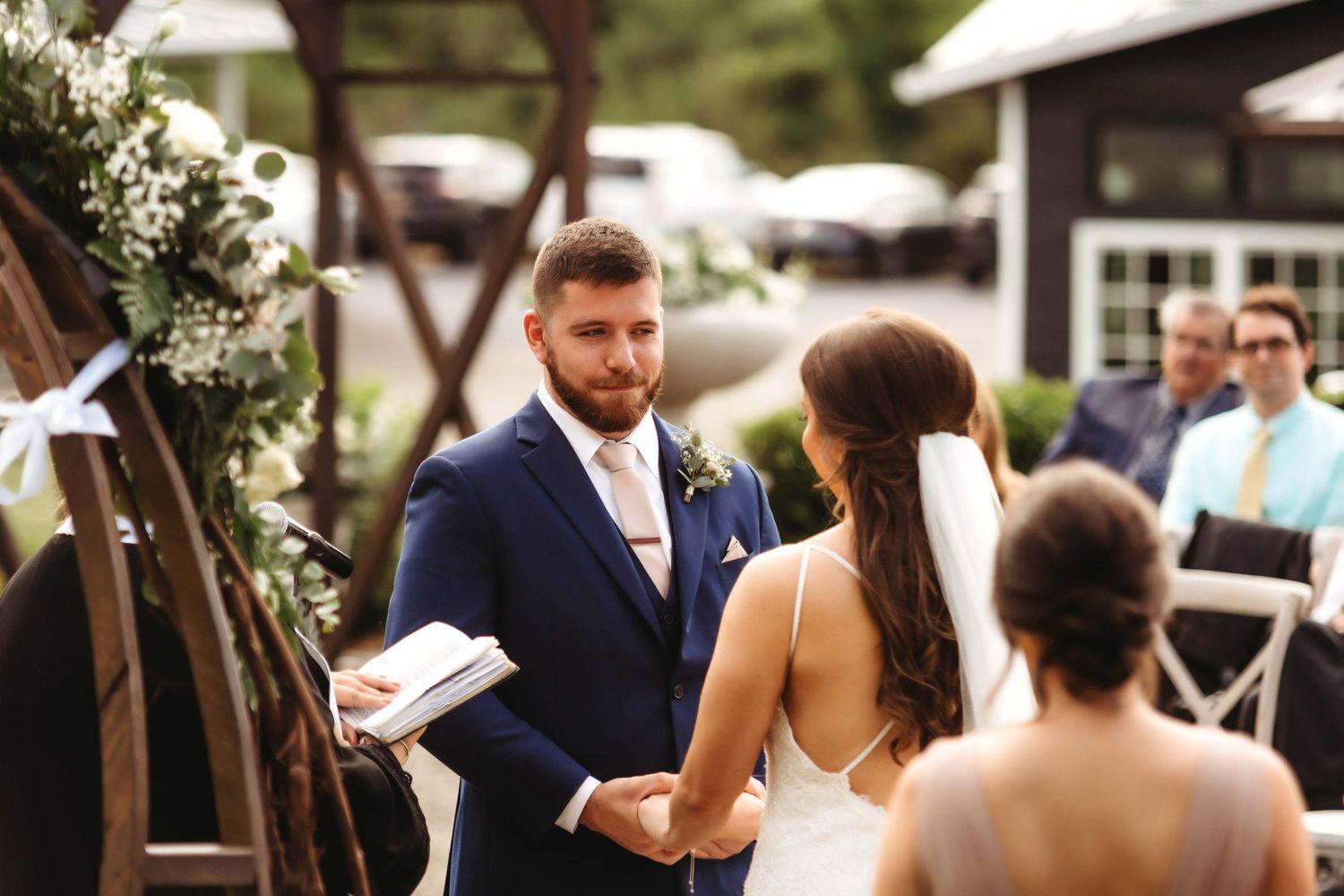 Guests gathered outdoors for a wedding ceremony beneath trees, with white umbrellas and ceremony chairs.