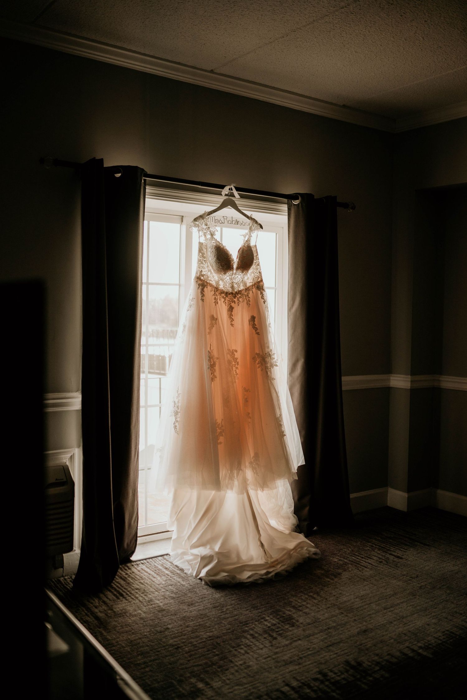 Ethereal wedding dress hanging in silhouette against a sunlit window in a dark room.