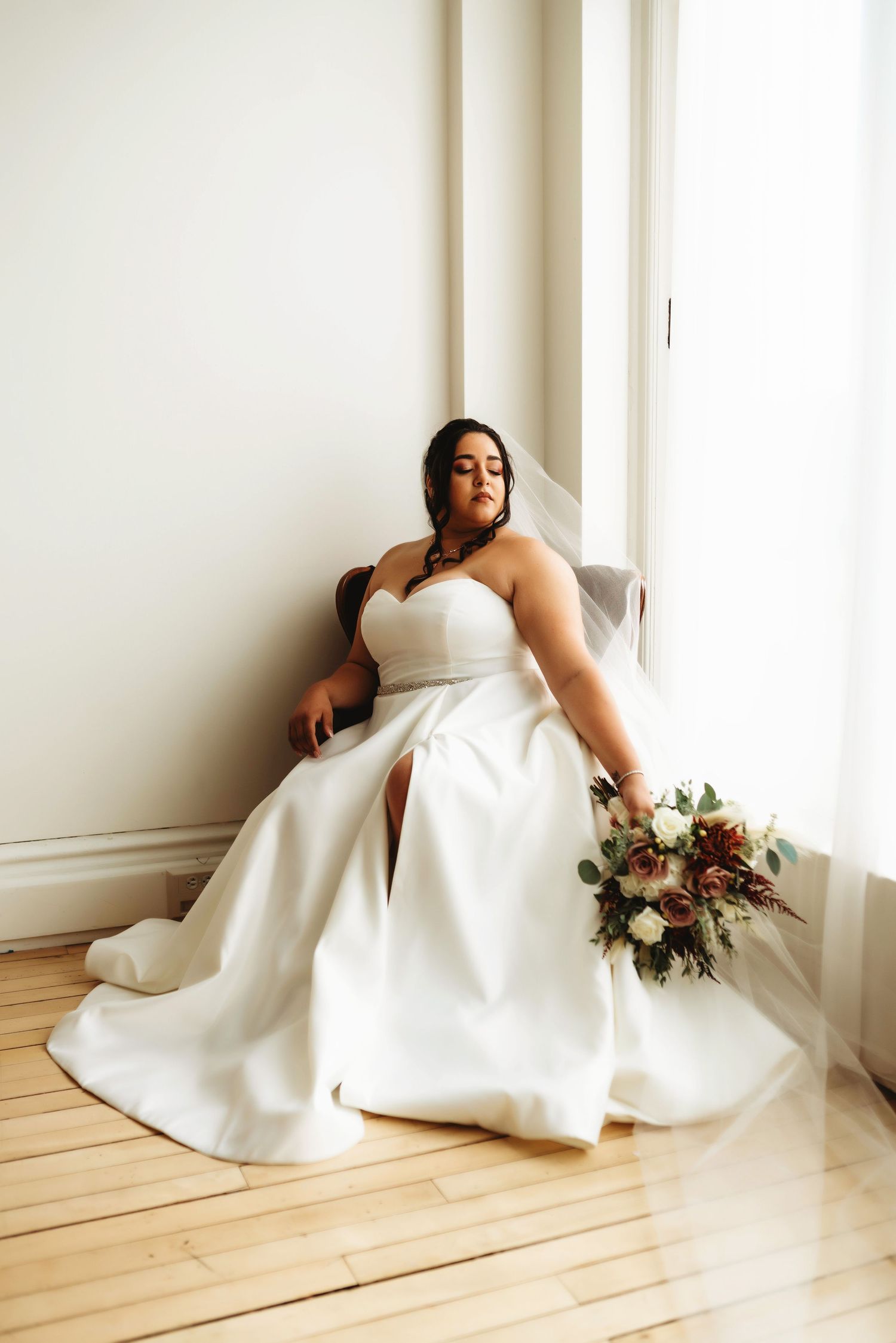 Elegant bridal portrait of a bride in a white gown seated near a window, photographed in soft natural light.