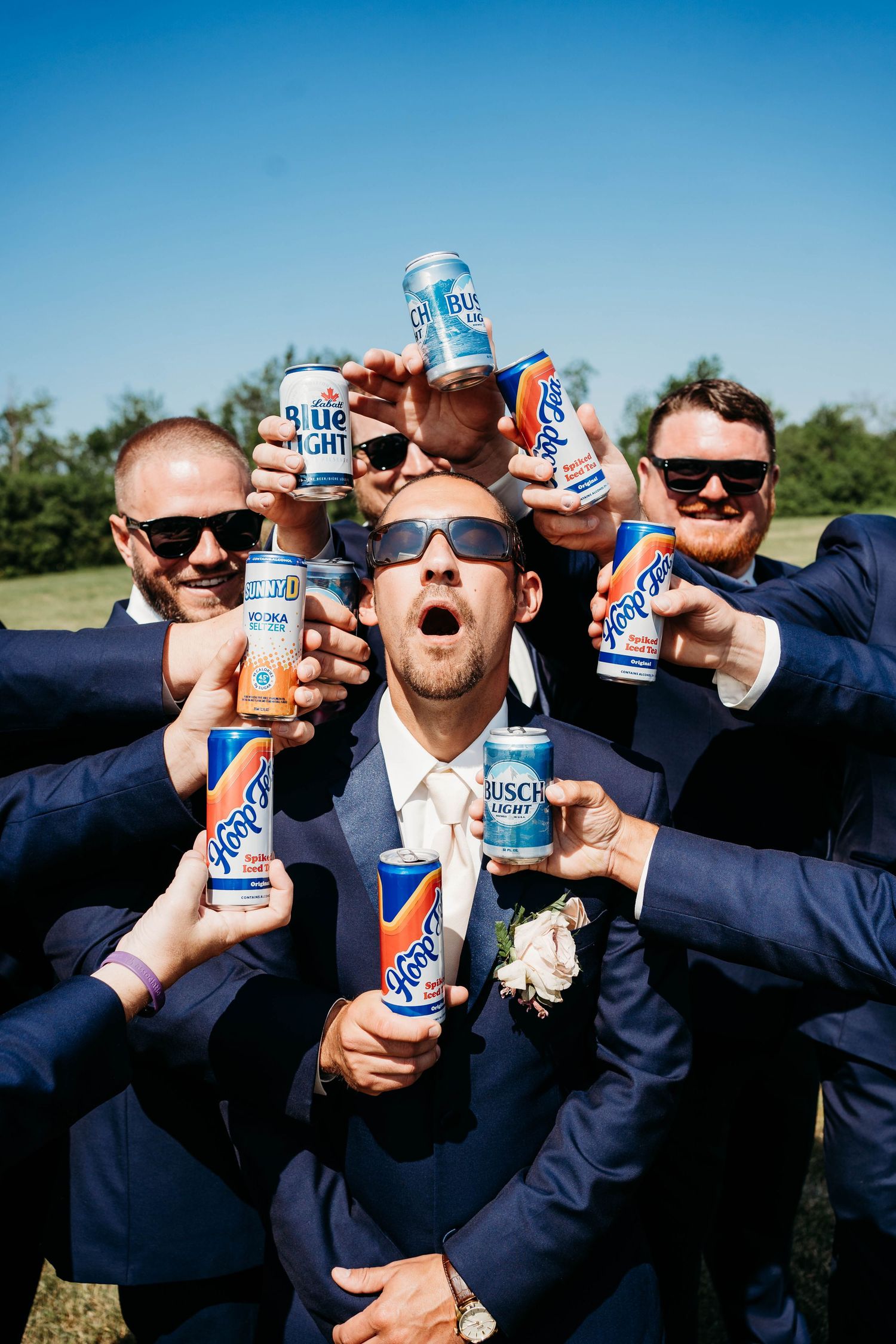 Groomsmen in navy suits holding up Busch Light beer cans in a celebratory pose outdoors on a sunny day.