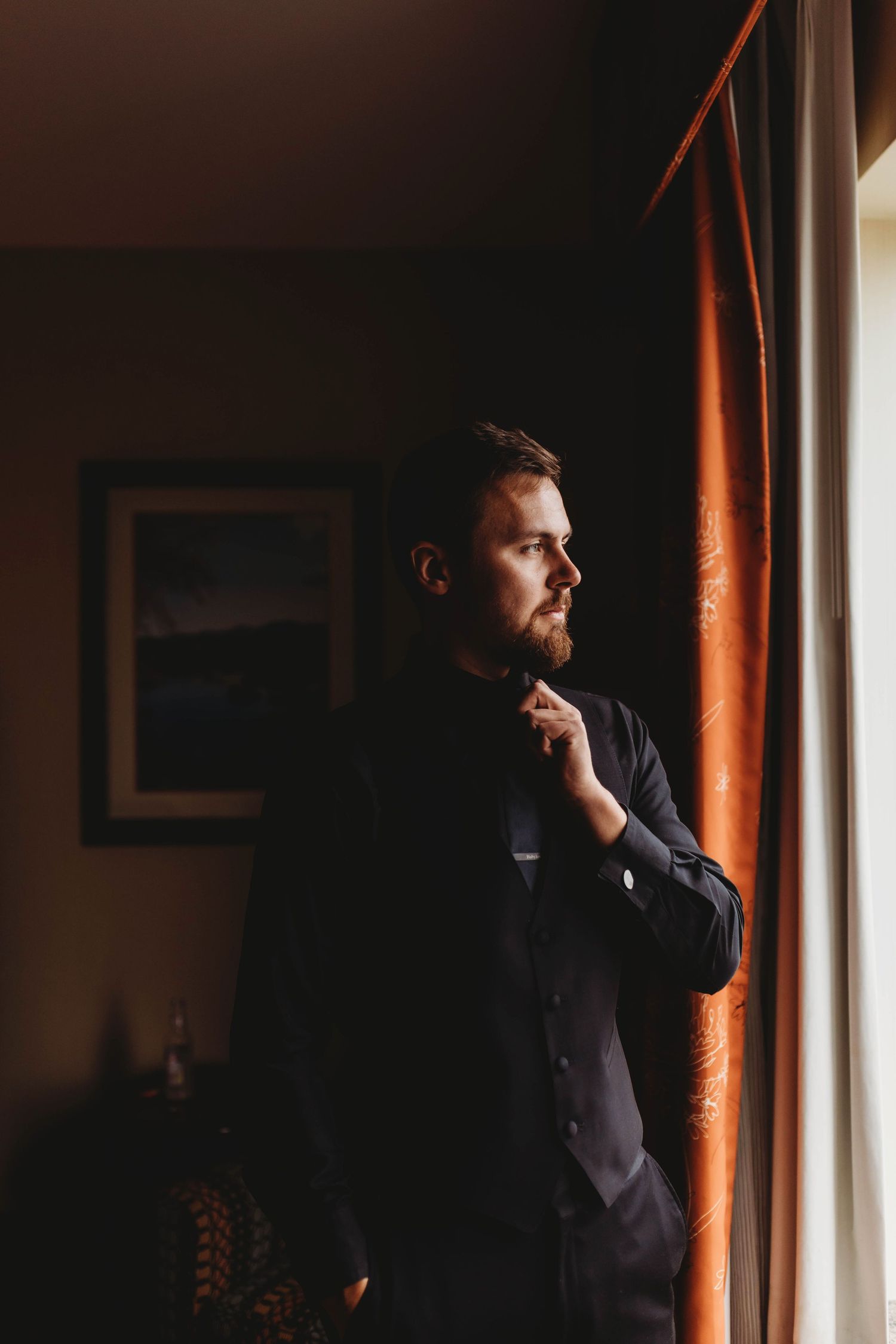 A portrait of the groom in dark clothing gazes thoughtfully through a window in warm, dramatic lighting.