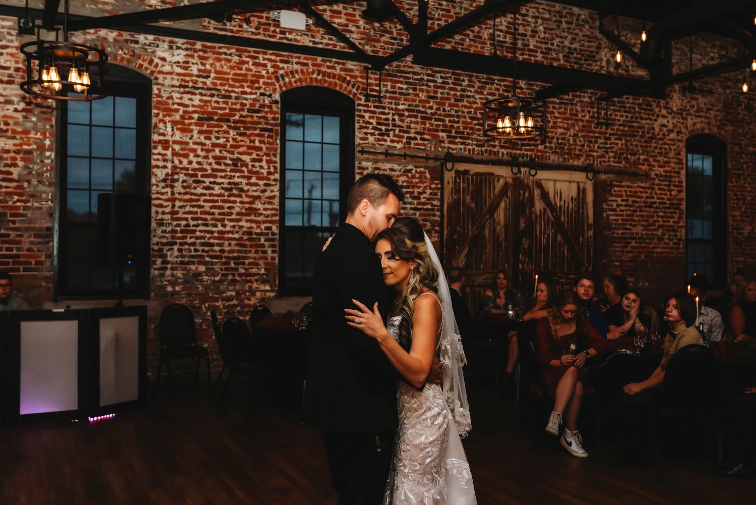 A wedding couple dances together during their first dance at a rustic brick venue with warm lighting and exposed beams. Bride and groom