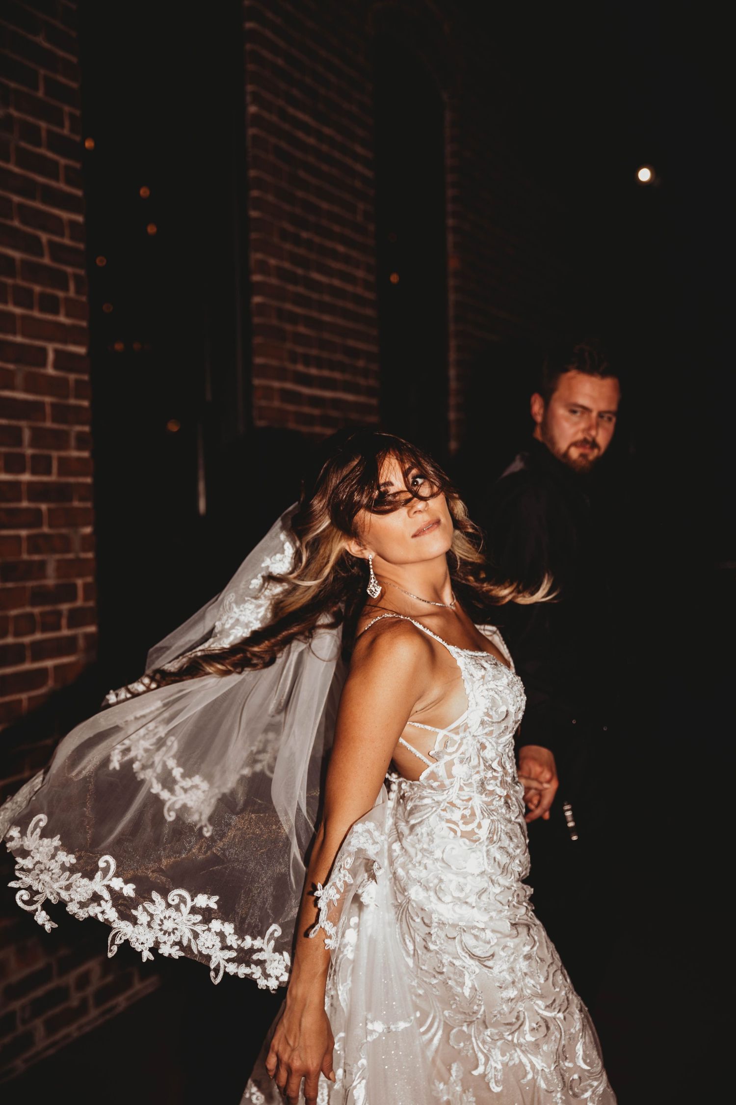 A bride in a white lace wedding dress with flowing veil dances joyfully against a dark brick wall backdrop.