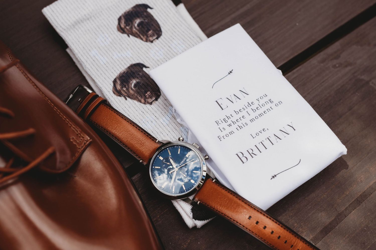 Wedding day flat lay featuring a groom's watch, leather accessories, and invitation suite arranged on a dark surface.