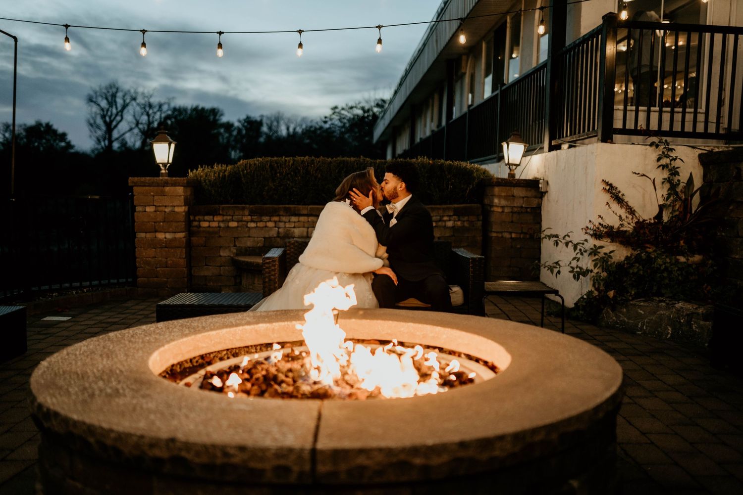 Bride and groom share an intimate moment- String lights illuminate an outdoor fire pit area with cozy seating and glowing flames at dusk.