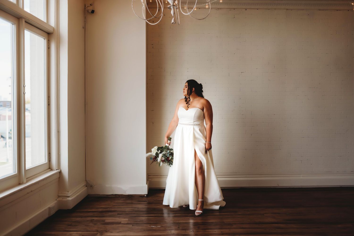 Bride in a white wedding dress standing by large windows in an elegant room with soft natural light.