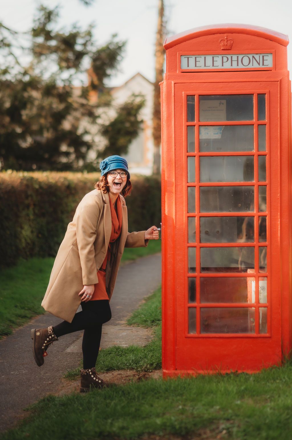Creative marketing consultant laughing beside a red telephone box, wearing glasses, a blue hat and a coat.
